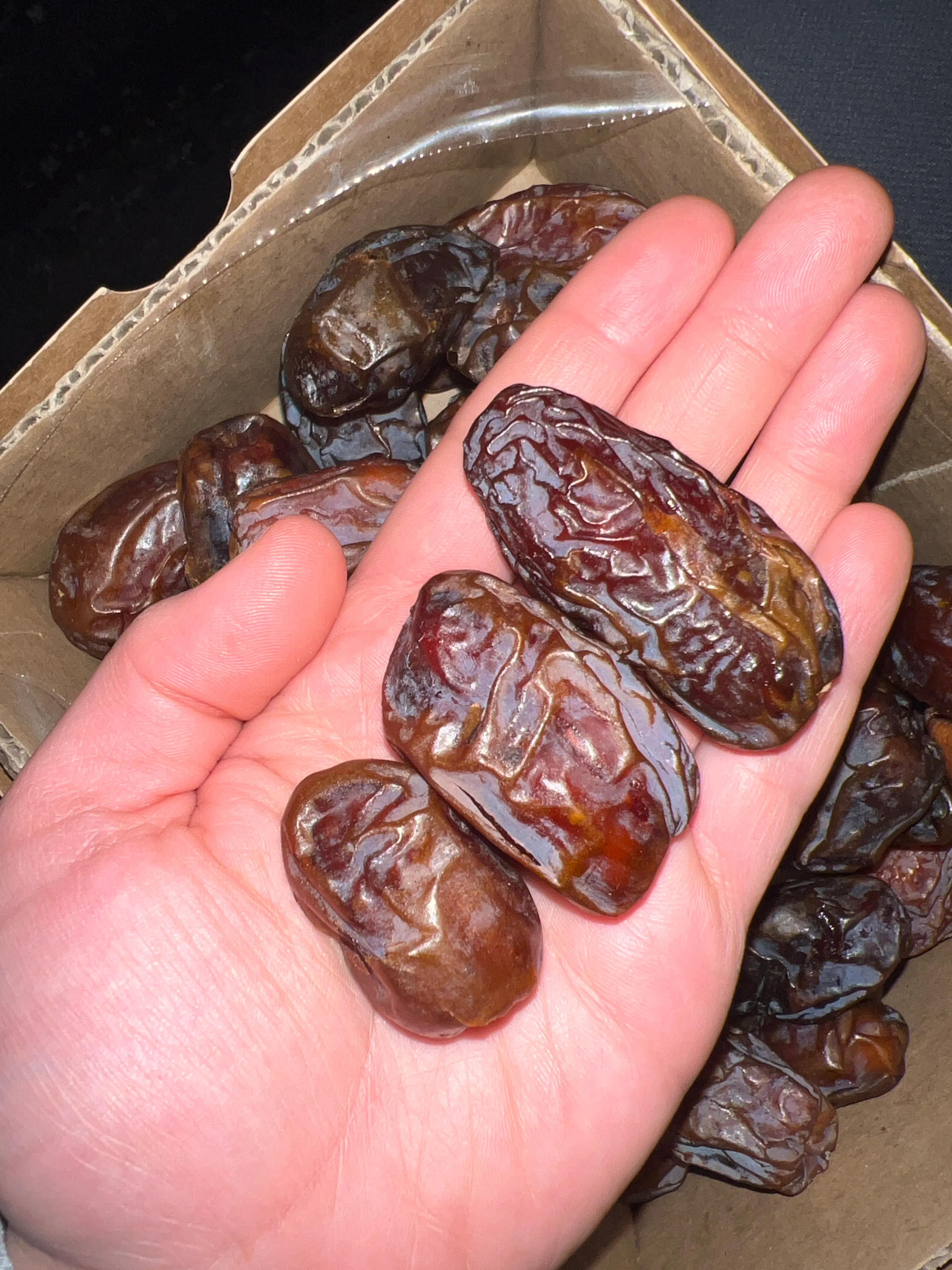 a hand holds several dates above a cardboard box filled with more dates.
