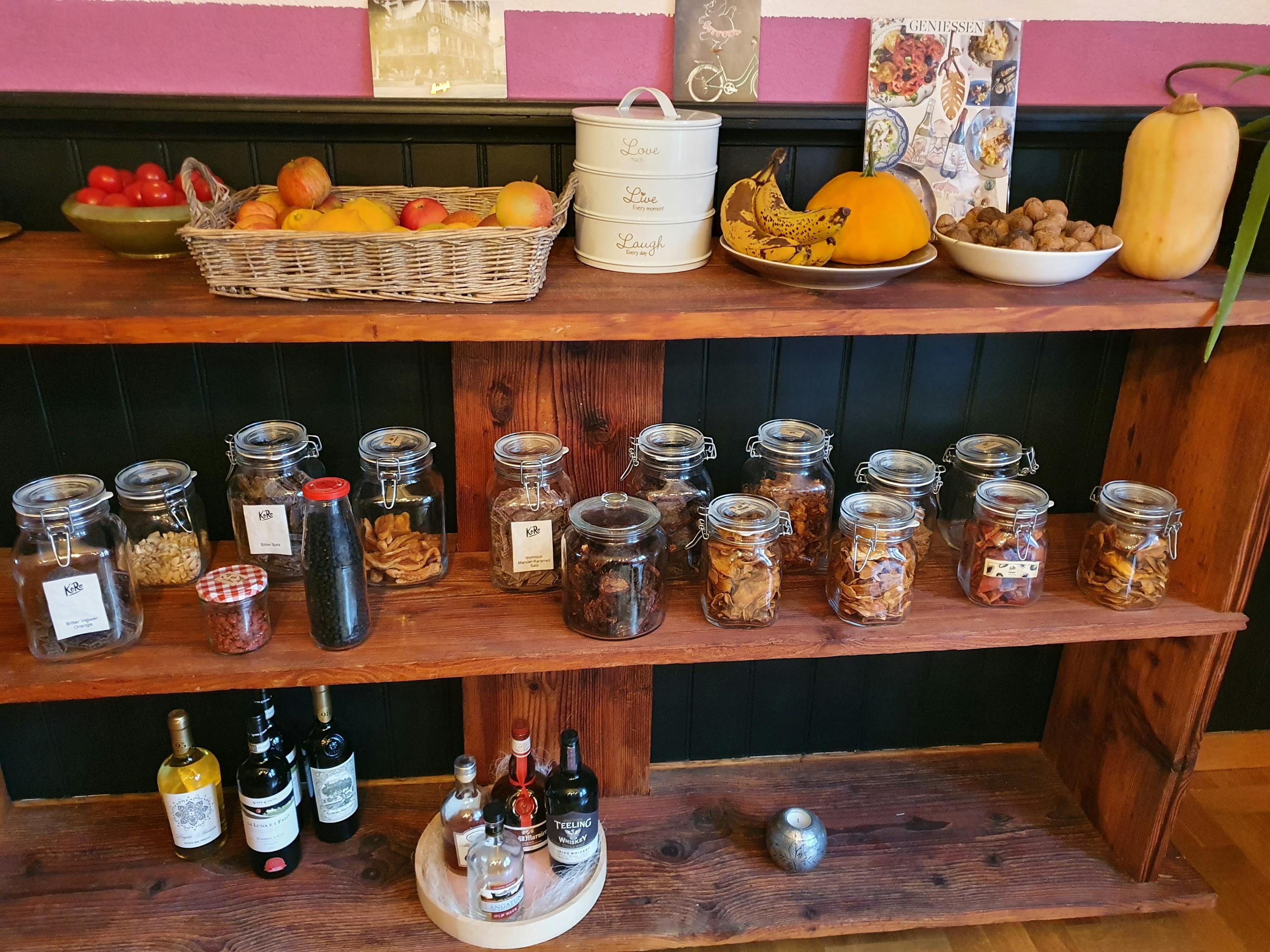 ein Holzregal mit Gläsern und Flaschen mit Etiketten auf den Gläsern steht neben einem Korb mit Früchten