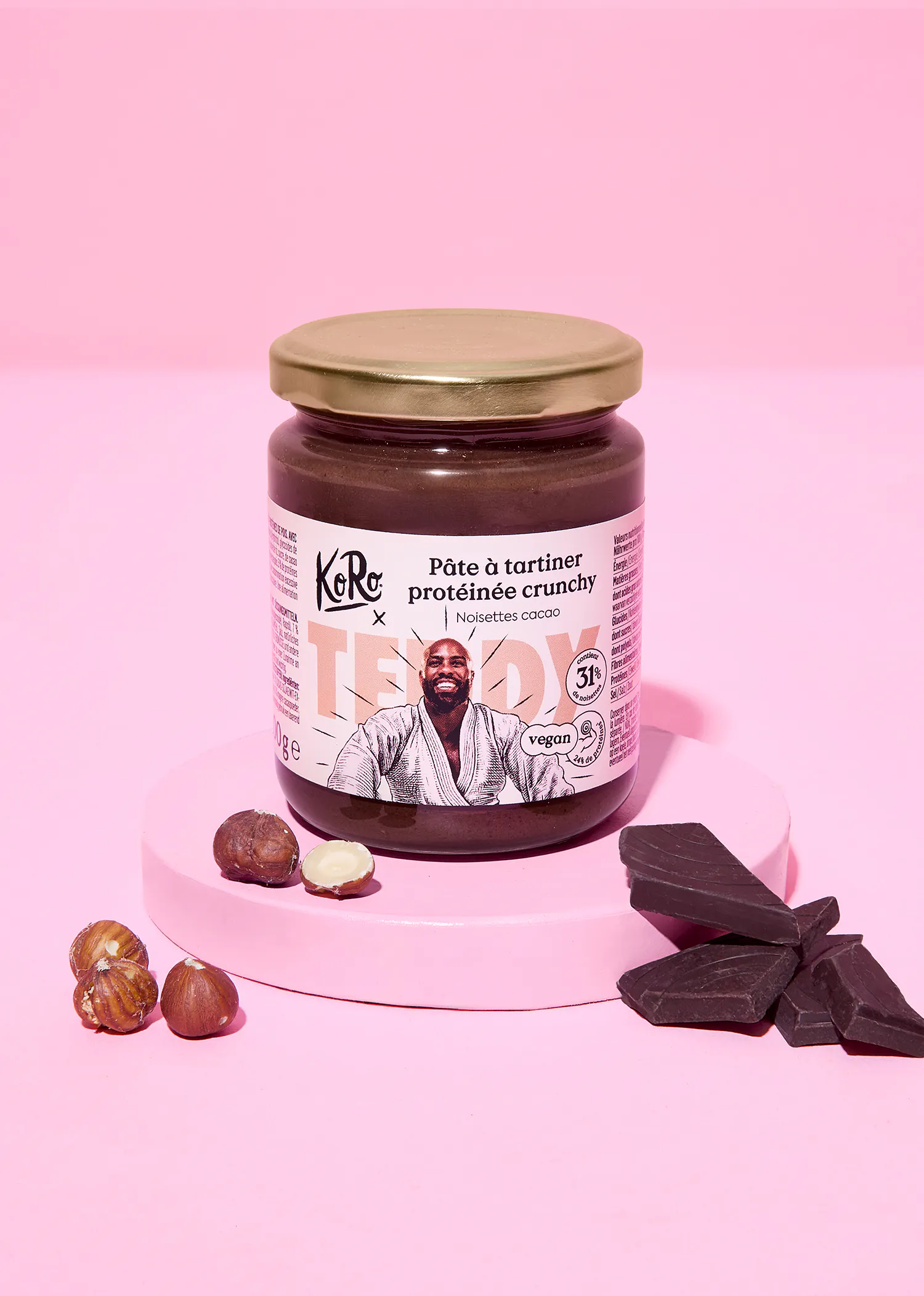 un pot de pâte à tartiner protéinée crunchy noisettes cacao koro x teddy, avec un homme souriant en kimono sur l'étiquette, posé sur un socle rose entouré de noisettes et de morceaux de chocolat noir, sur un fond rose uni.
