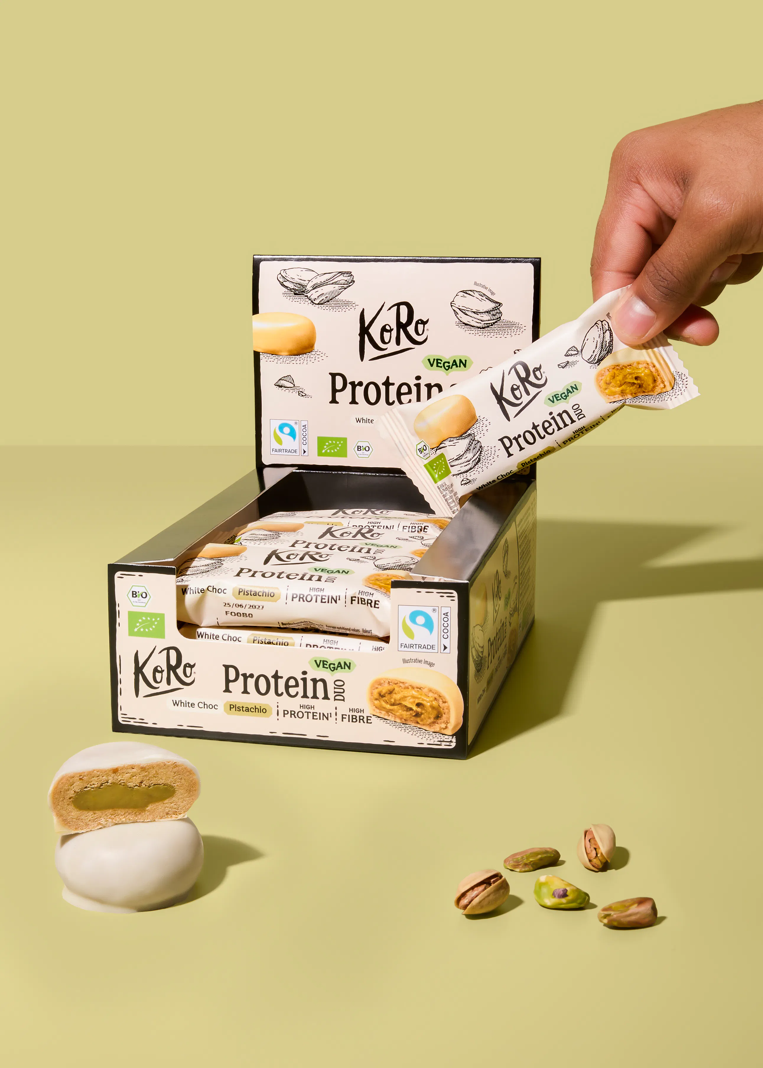 eine hand nimmt einen weißen schoko-pistazien-proteinriegel von koro aus einer geöffneten schachtel, während ein weiterer riegel aufgeschnitten seine grüne füllung zeigt und lose pistazien daneben liegen.
