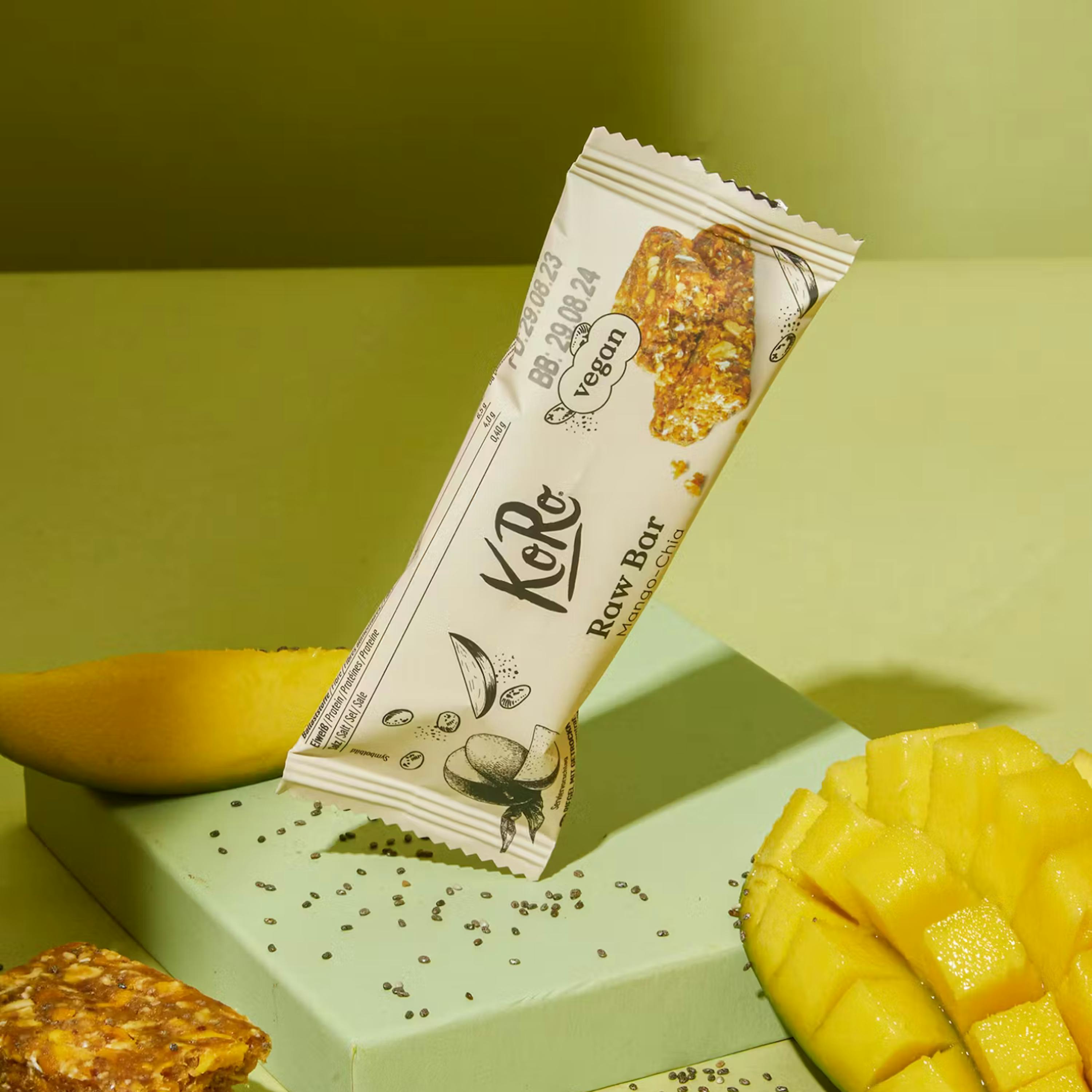 una barretta cruda koro mango-chia in piedi su un blocco verde chiaro, circondata da fette di mango e semi di chia.