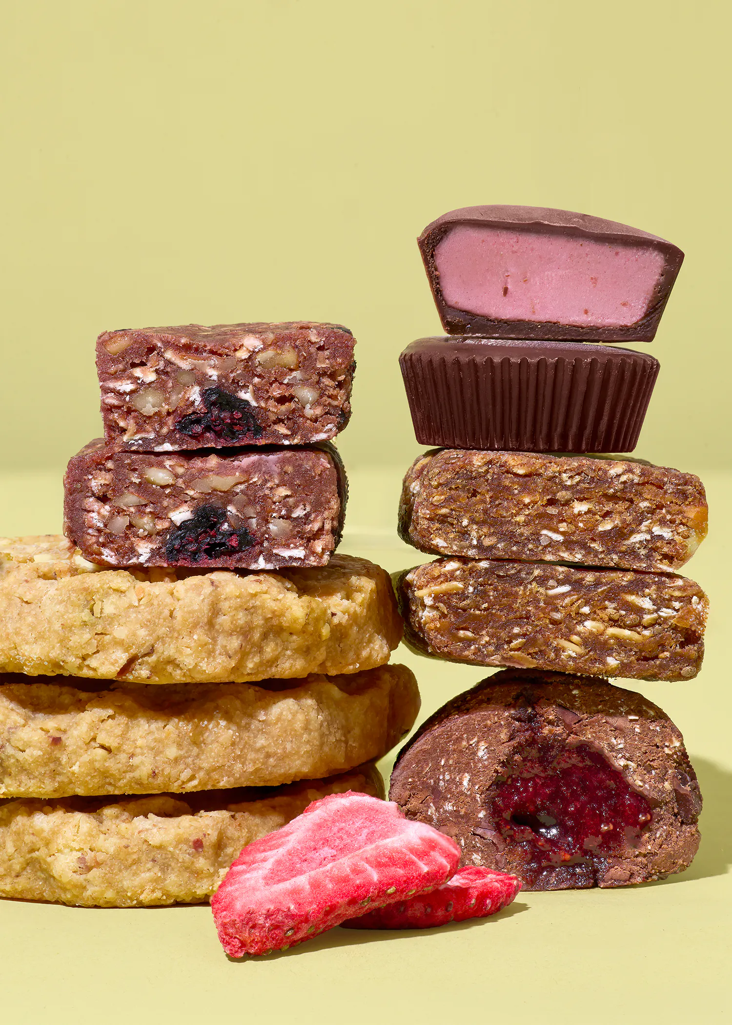 eine anordnung von gestapelten süßen snacks, darunter nussriegel, kekse, schokoladenförmchen mit rosa füllung und ein schokoladensnack mit roter füllung, zusammen mit gefriergetrockneten erdbeeren auf hellgelbem hintergrund.