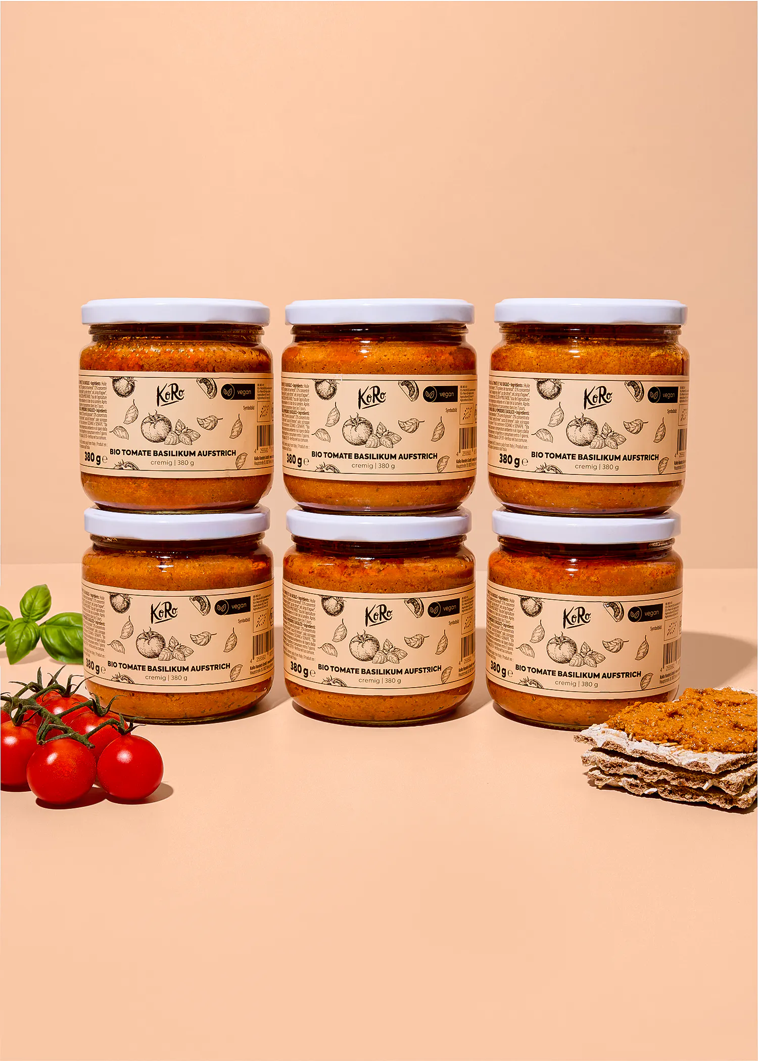 sei barattoli di crema spalmabile koro al pomodoro e basilico disposti su due file su uno sfondo color pesca, accompagnati da pomodorini freschi e una fetta di pane croccante.