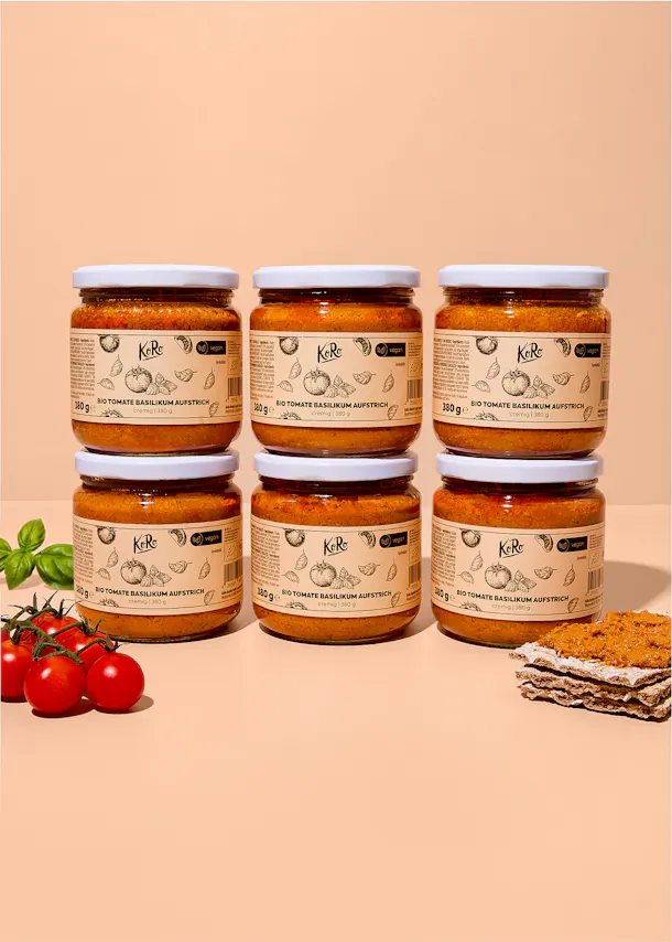 sei barattoli di crema spalmabile koro al pomodoro e basilico disposti su due file su uno sfondo color pesca, accompagnati da pomodorini freschi e una fetta di pane croccante.
