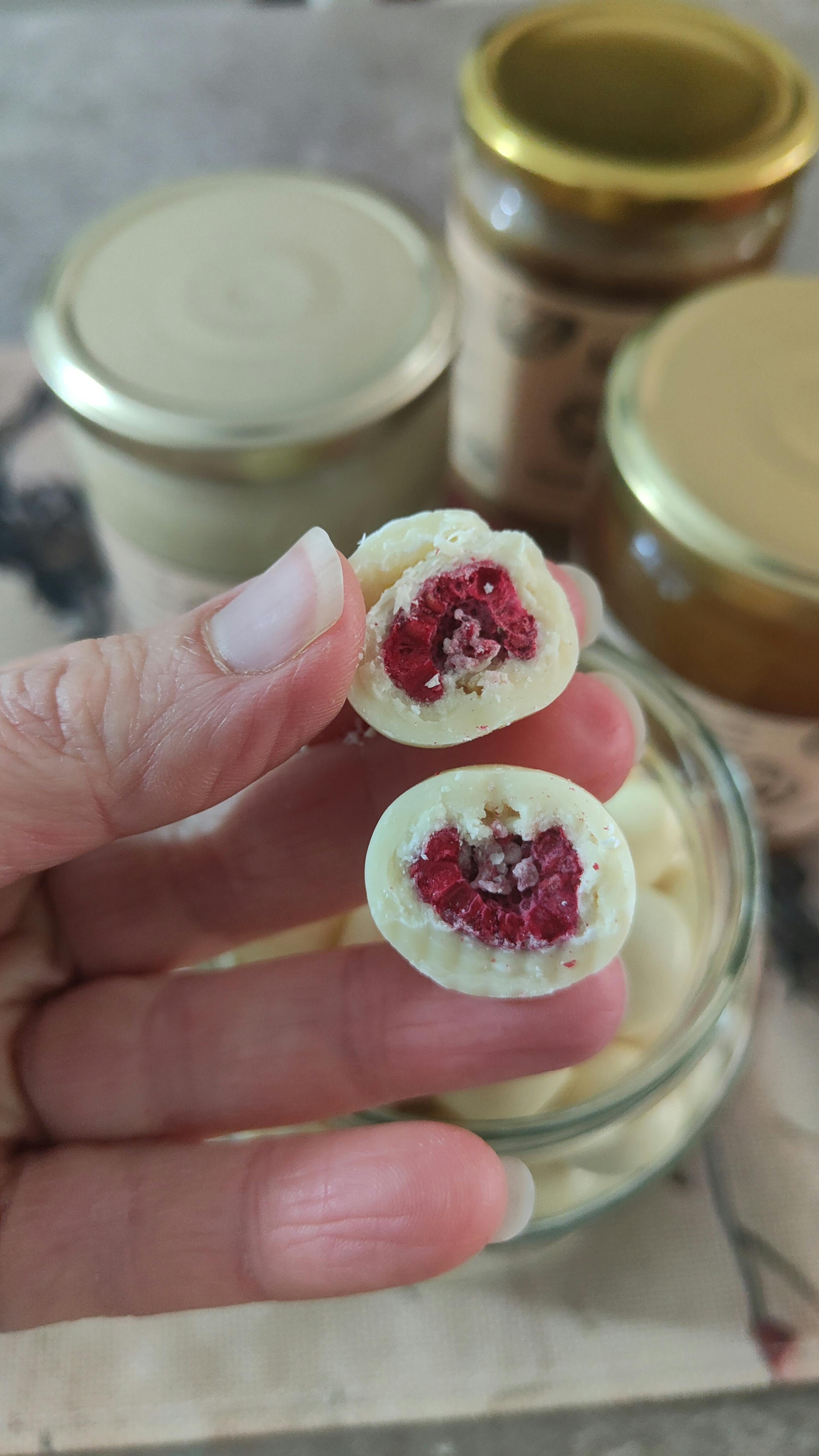 eine hand hält zwei halbierte, weiß schokolierte himbeeren, die ihr rotes inneres zeigen, mit weiteren gläsern im hintergrund.