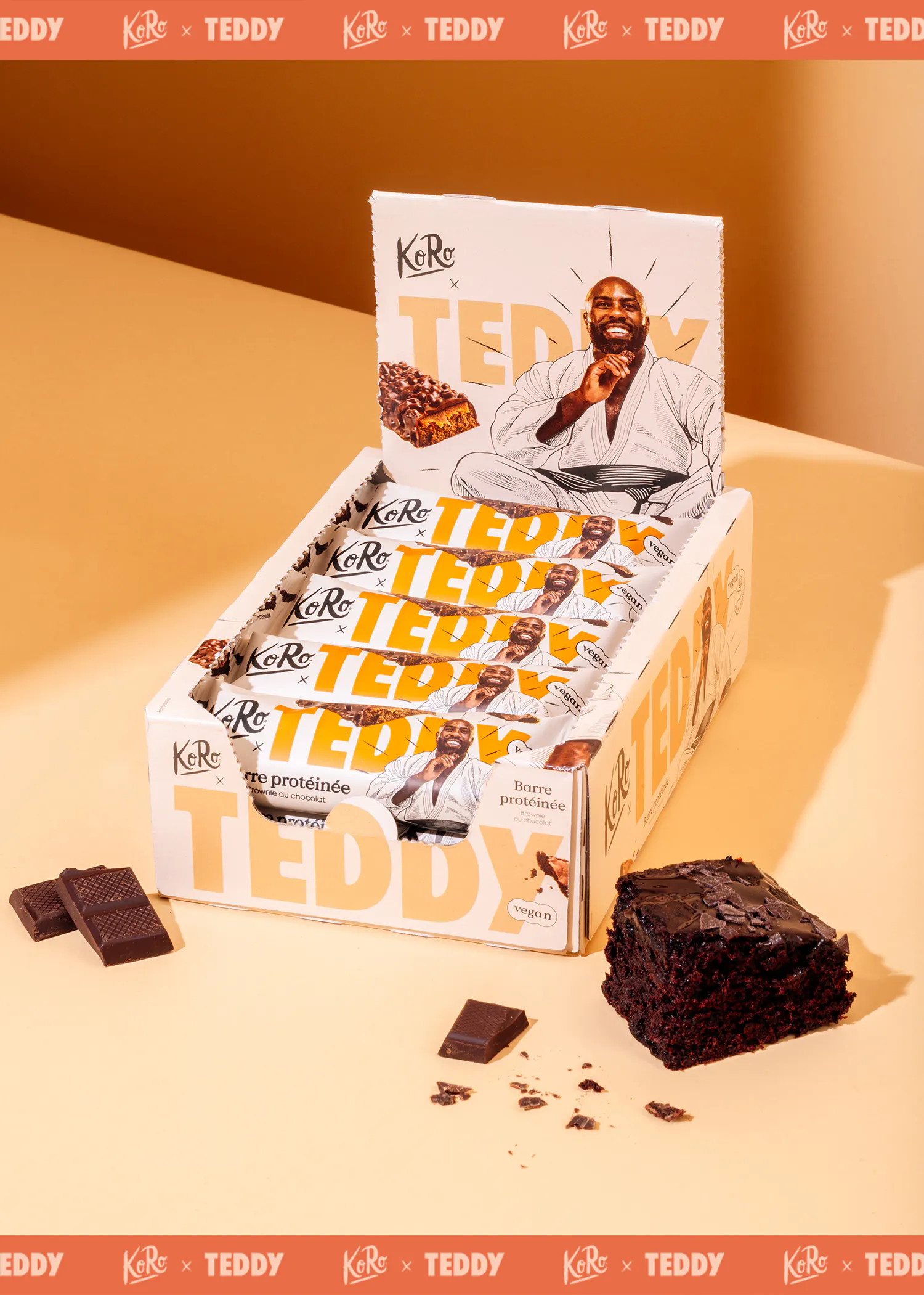 une boîte de présentation de barres protéinées véganes koro x teddy saveur brownie au chocolat, avec un homme souriant en kimono sur l'emballage, accompagnée d'un morceau de brownie et de carrés de chocolat.