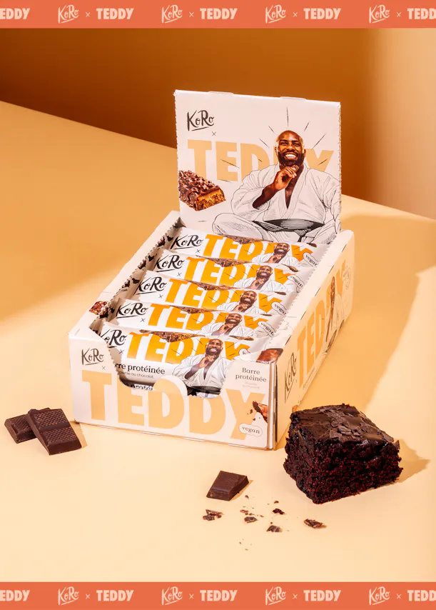 une boîte de présentation de barres protéinées véganes koro x teddy saveur brownie au chocolat, avec un homme souriant en kimono sur l'emballage, accompagnée d'un morceau de brownie et de carrés de chocolat.