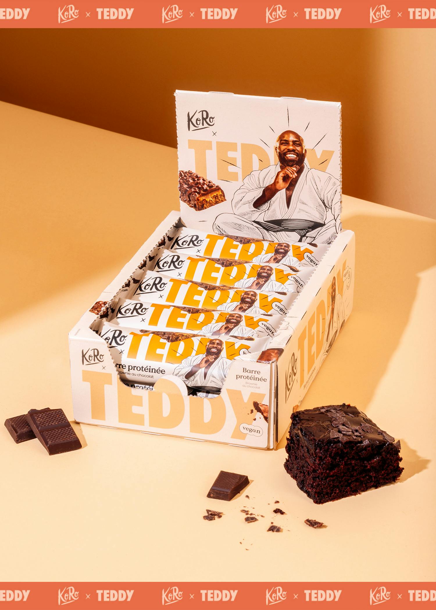 Barre protéinée Teddy vegan au chocolat brownie 15 x 55 g une boîte de présentation de barres protéinées véganes koro x teddy saveur brownie au chocolat, avec un homme souriant en kimono sur l'emballage, accompagnée d'un morceau de brownie et de carrés de chocolat.