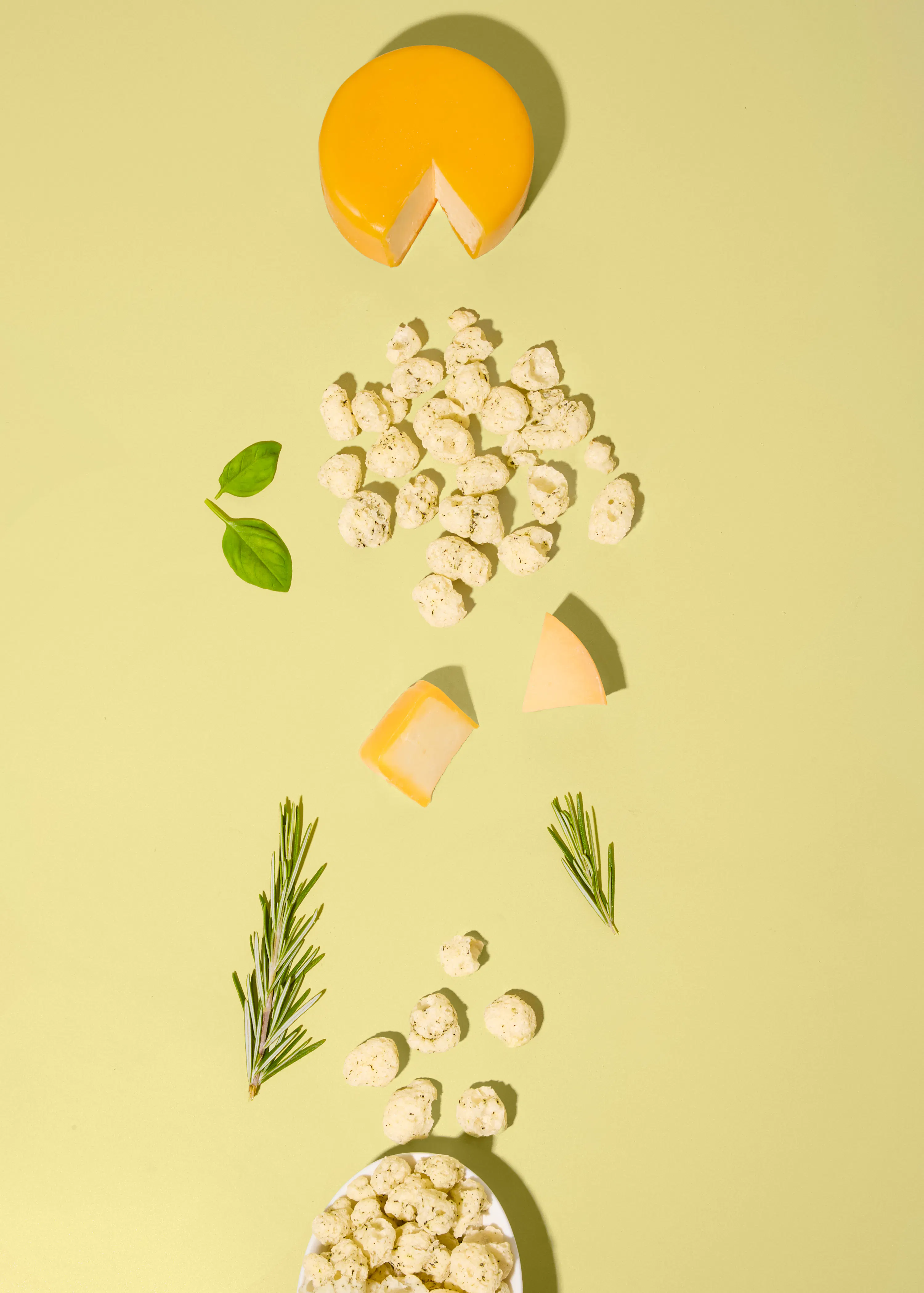 una composizione dall'alto di formaggio a pasta dura, snack croccanti, foglie di basilico e rametti di rosmarino disposti su uno sfondo verde chiaro.