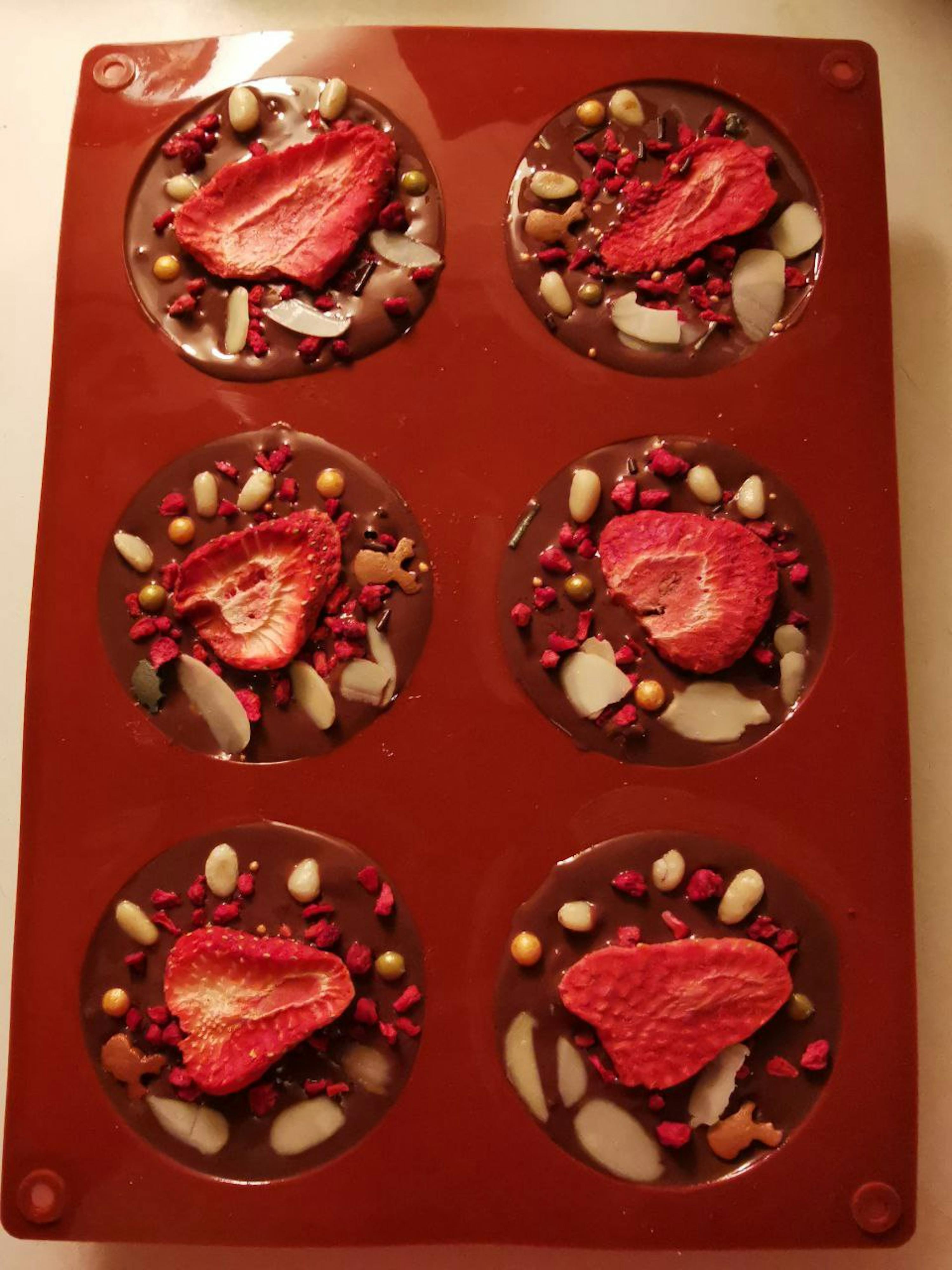 a red silicone mold contains six circular chocolate candies, each adorned with a dried strawberry slice, red and gold sprinkles, and almond slivers.