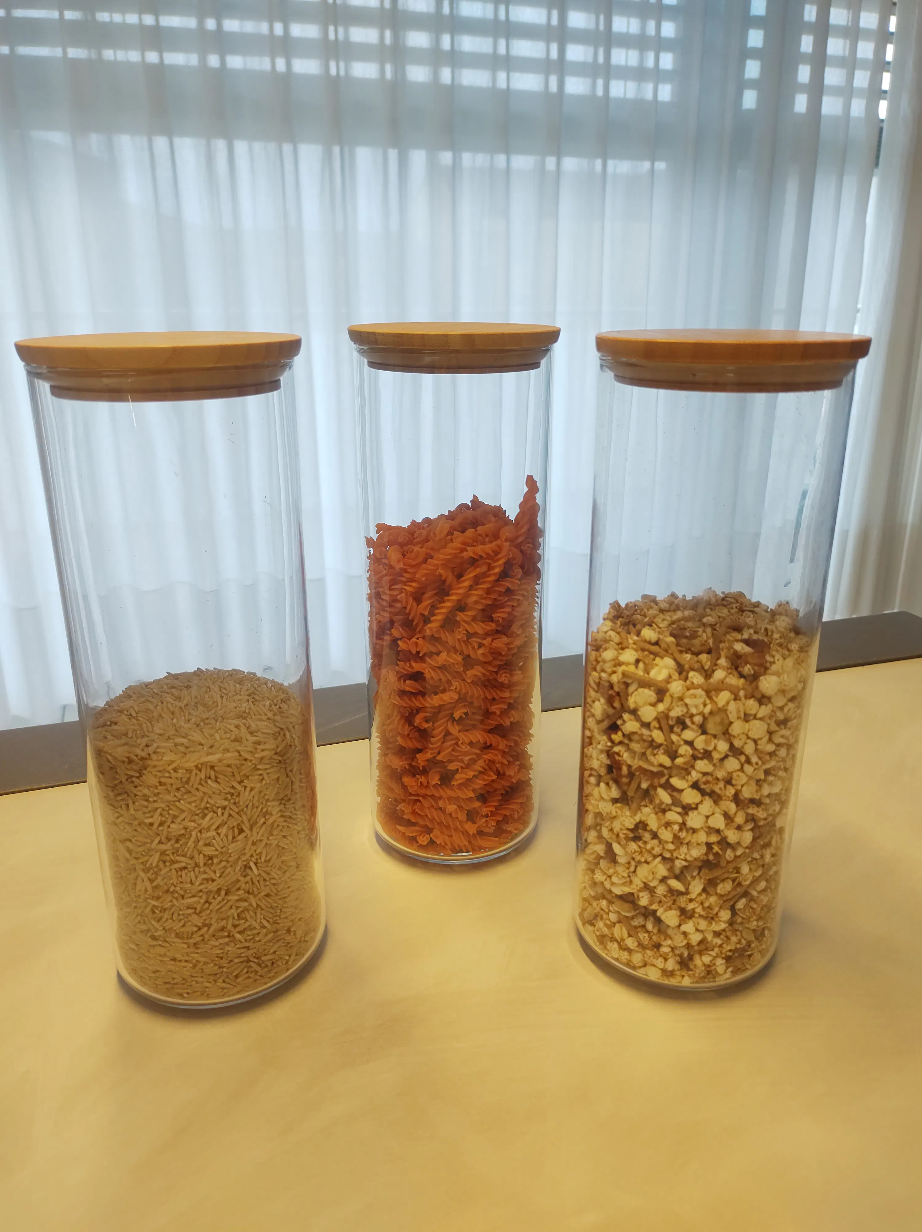 drei transparente glasgefäße mit holzdeckeln, gefüllt mit reis, roter fusilli-pasta und müsli, stehen vertikal vor einem hellen hintergrund.