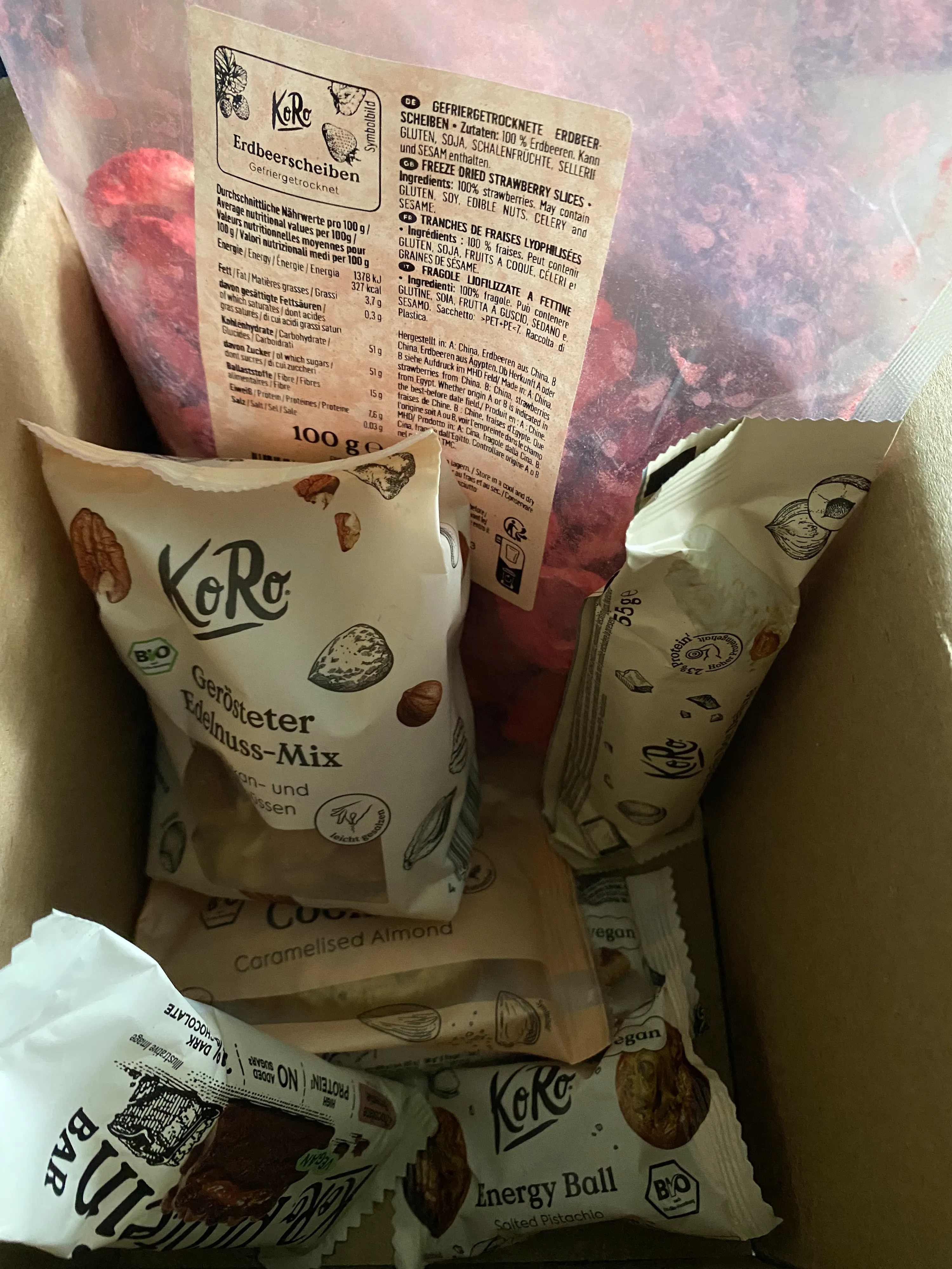 eine offene versandbox ist gefüllt mit verschiedenen snackpackungen, darunter gefriergetrocknete erdbeerscheiben, nussmischungen, energiekugeln und proteinriegel.