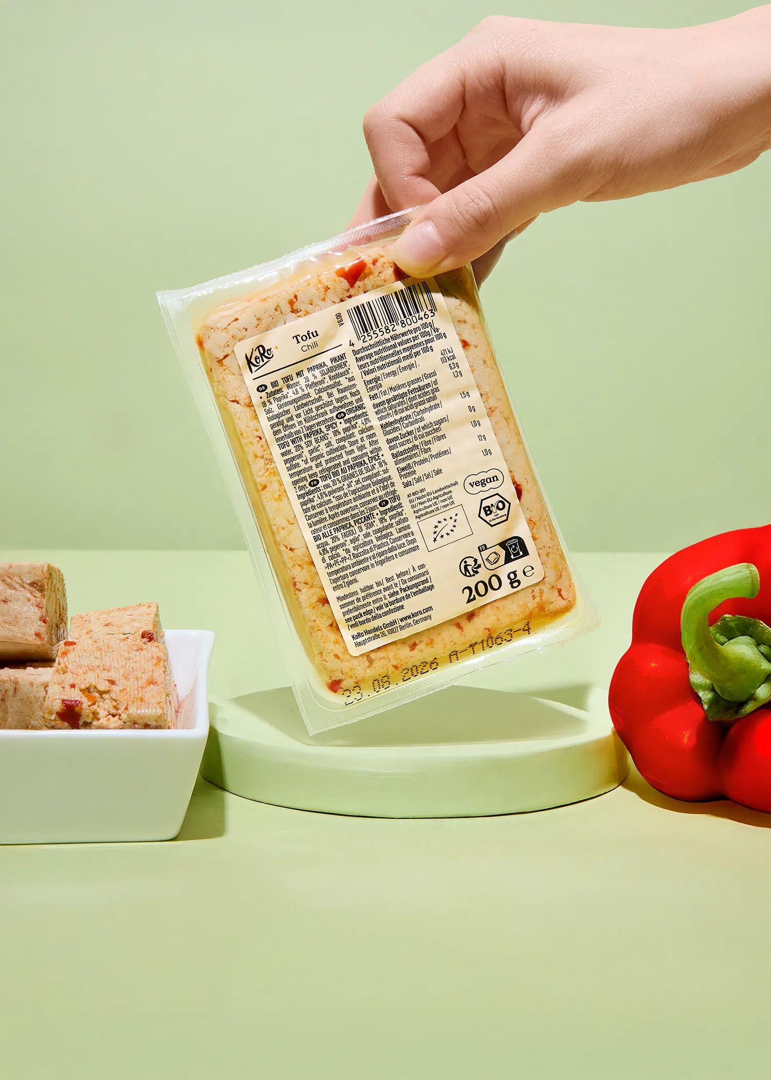 a hand holds a koro chili tofu package, with more tofu and a red bell pepper in the background.