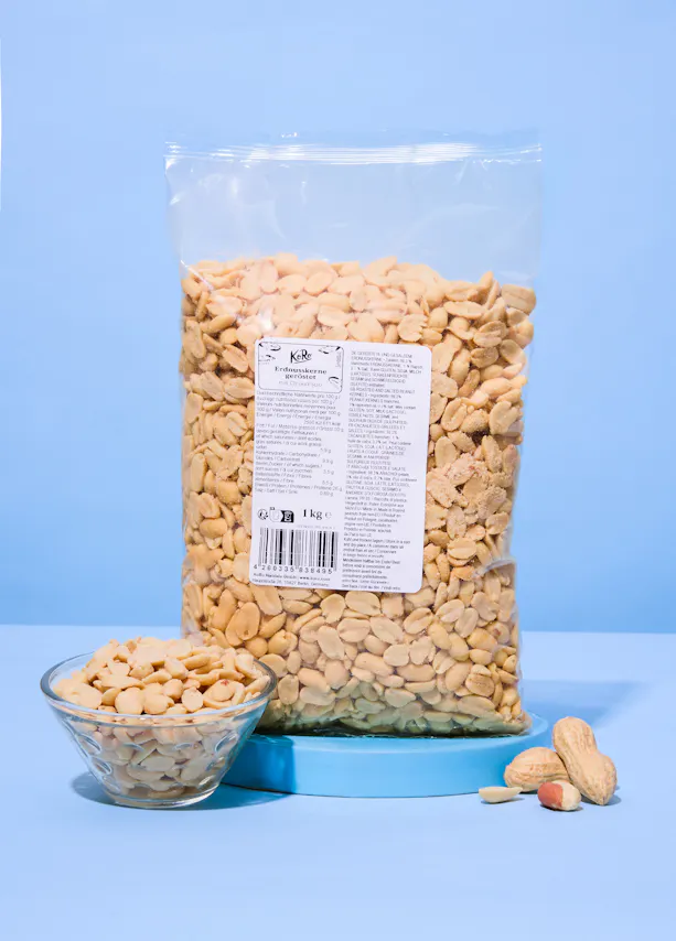 un grand sac transparent de cacahuètes grillées dorées de 1 kg avec une étiquette blanche, posé sur une surface bleue, accompagné d'un petit bol de cacahuètes et de quelques cacahuètes éparpillées au premier plan.