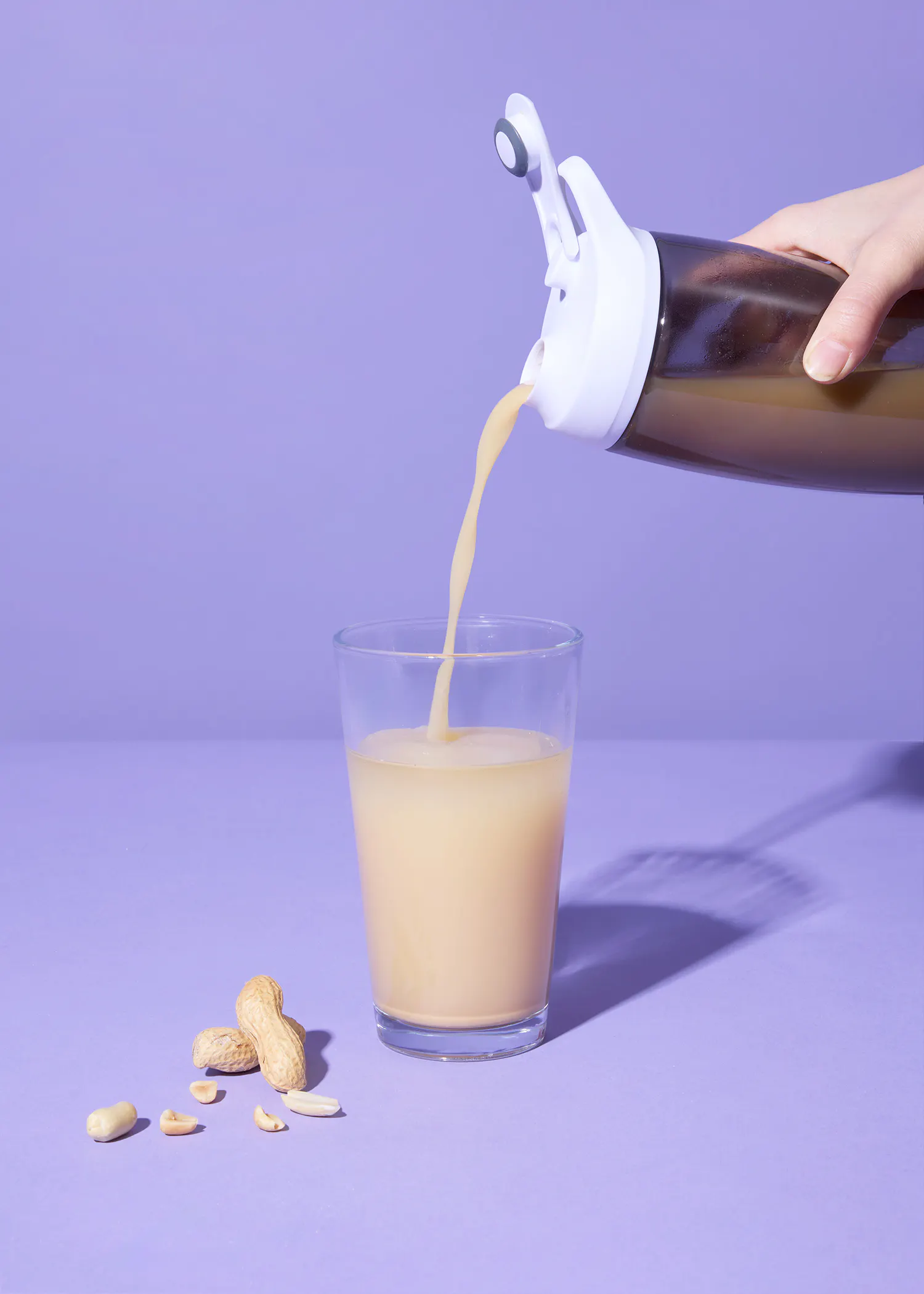 une main verse un liquide brun clair d'une bouteille dans un verre, avec des cacahuètes éparpillées à proximité sur un fond violet.