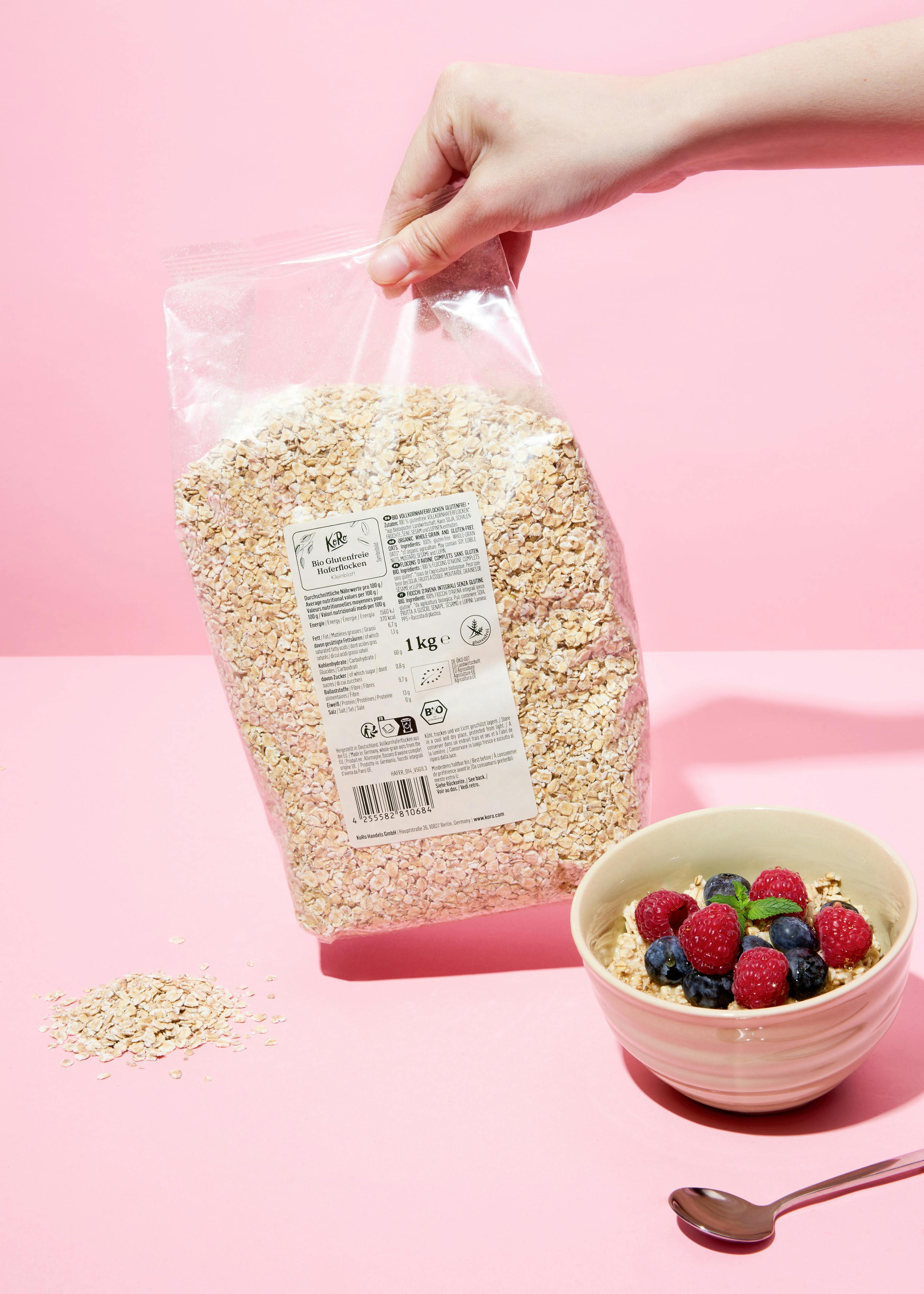 a hand holds a clear bag of organic gluten-free oats next to a bowl of oatmeal topped with raspberries, blueberries, and a mint leaf, all on a pink background.