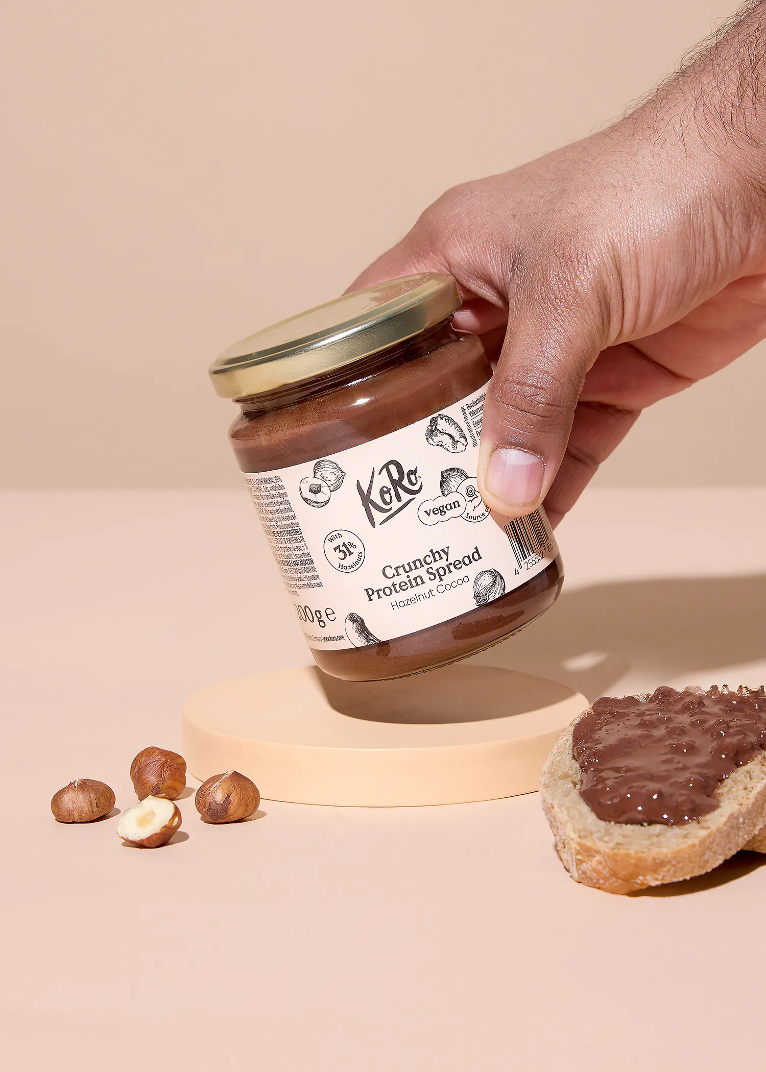 une main tient un pot de pâte à tartiner protéinée croquante koro noisette cacao, avec des noisettes et une tranche de pain tartinée visibles sur une surface beige clair.