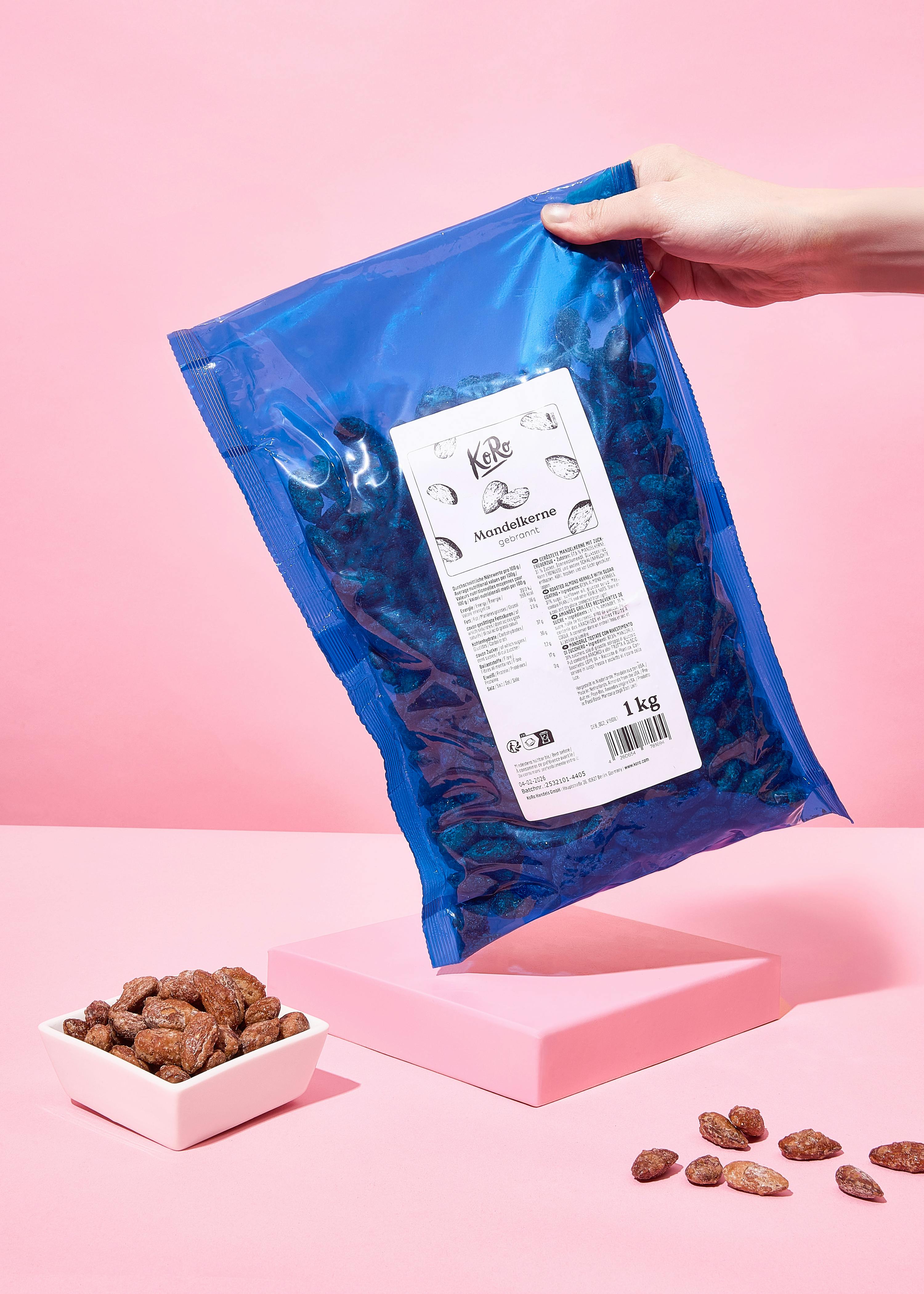 una mano tiene una busta blu da 1 kg di mandorle tostate, con una ciotola bianca e alcune mandorle sparse su una superficie rosa.