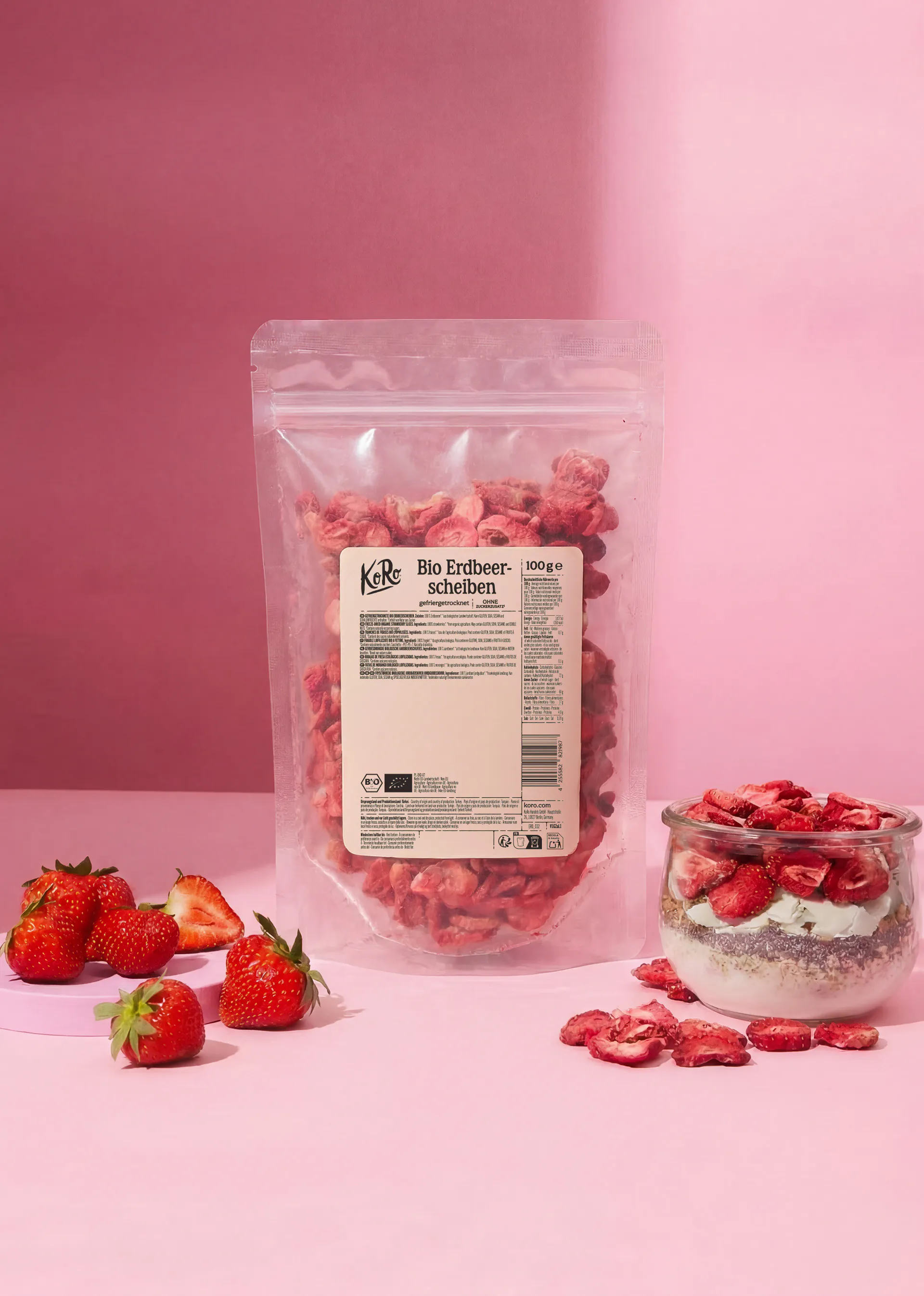 a clear pouch of koro organic freeze-dried strawberry slices stands next to a glass jar of yogurt topped with the same strawberries and a small pile of fresh strawberries on a pink background.