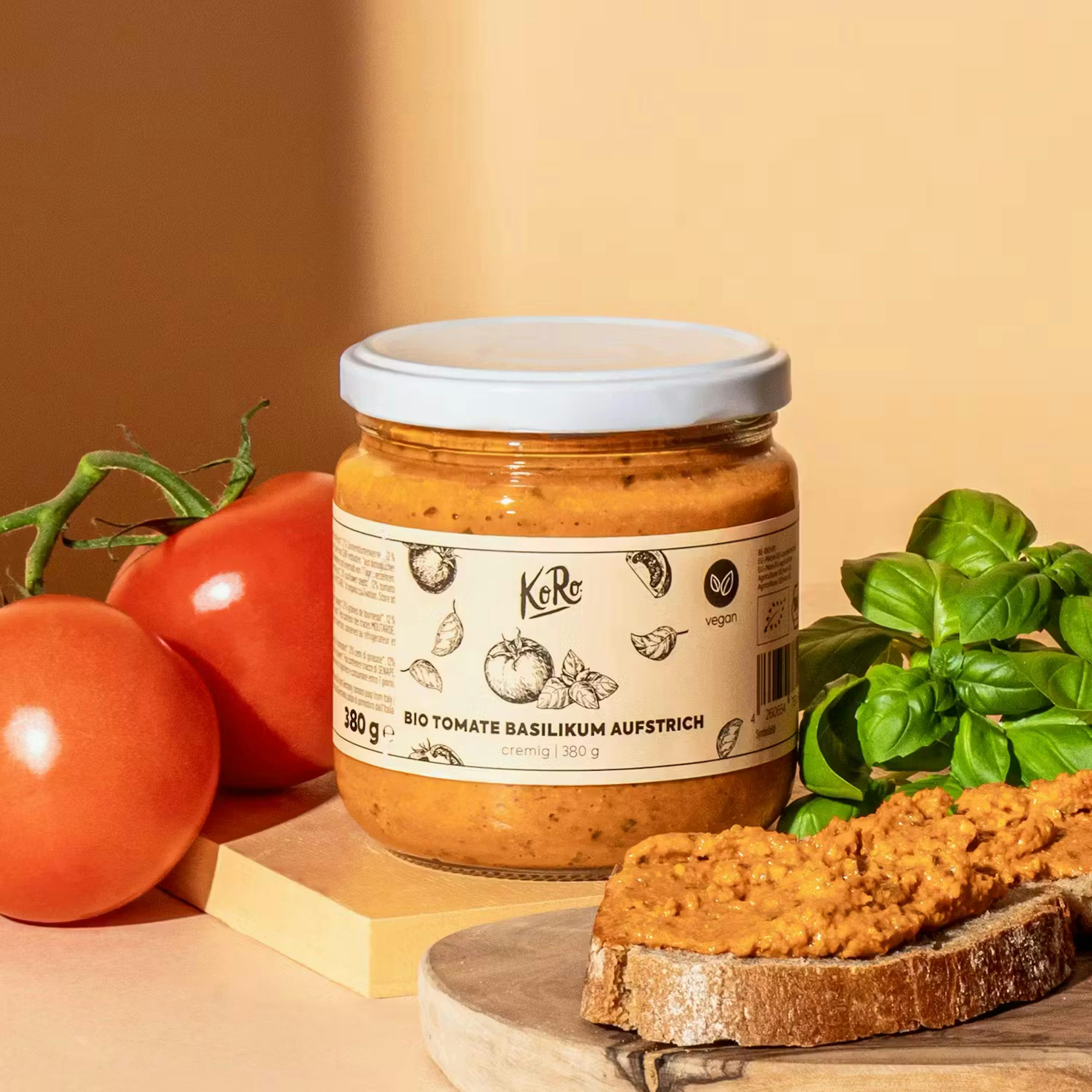 un pot de pâte à tartiner bio tomate basilic koro de 380g est entouré de tomates fraîches, de feuilles de basilic et d'une tranche de pain tartinée.