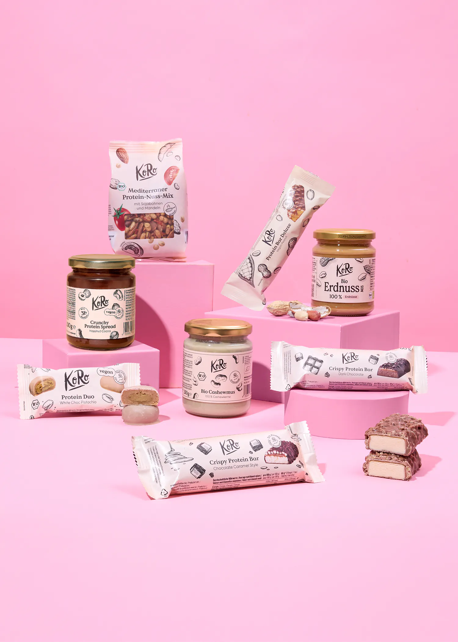 une variété de produits protéinés koro, incluant des pâtes à tartiner, des barres et un mélange de noix, sont présentés sur des blocs roses sur un fond rose.