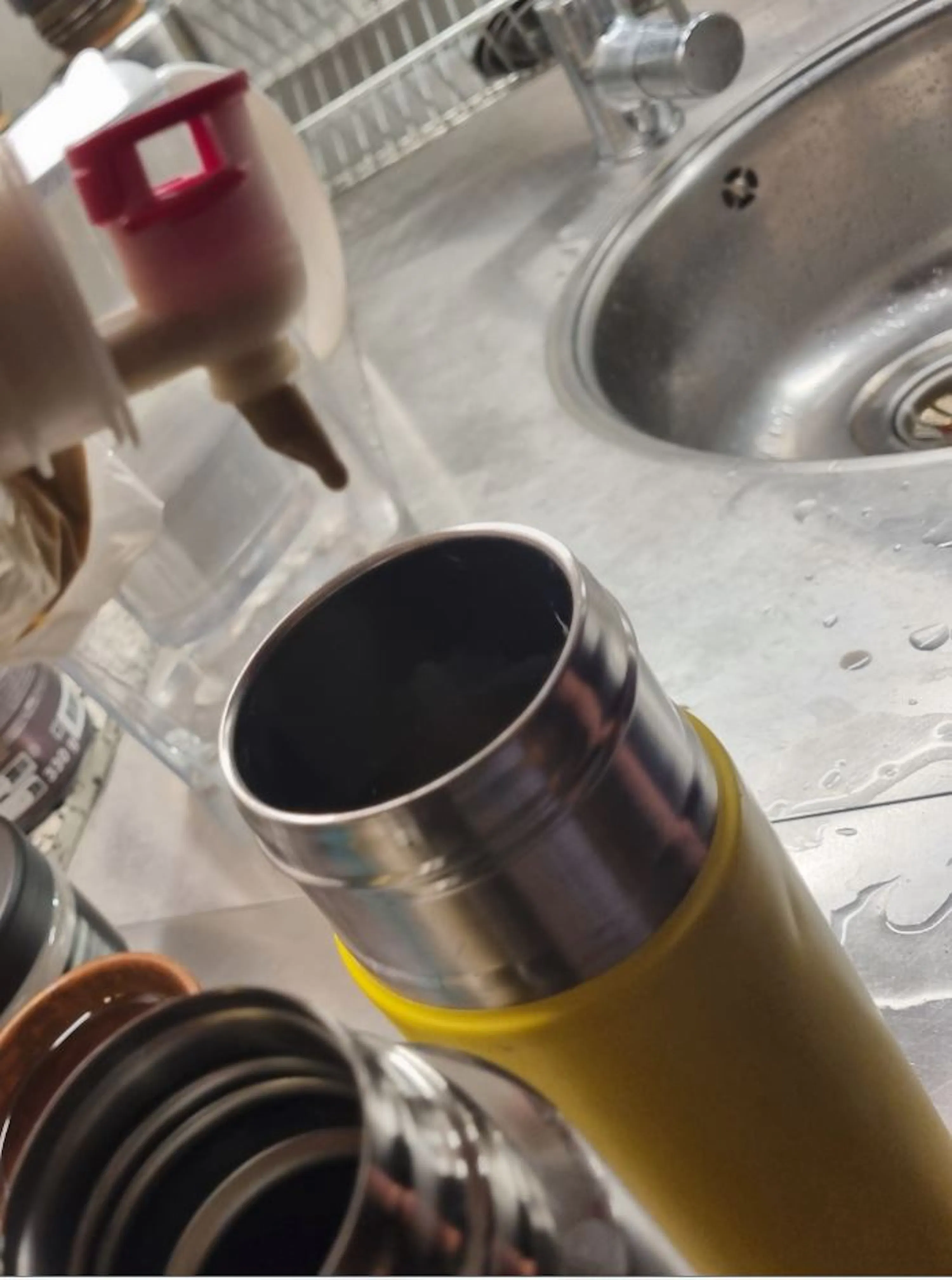 une bouteille thermos jaune ouverte repose sur un comptoir de cuisine humide à côté d'un évier, avec un biberon visible en arrière-plan.