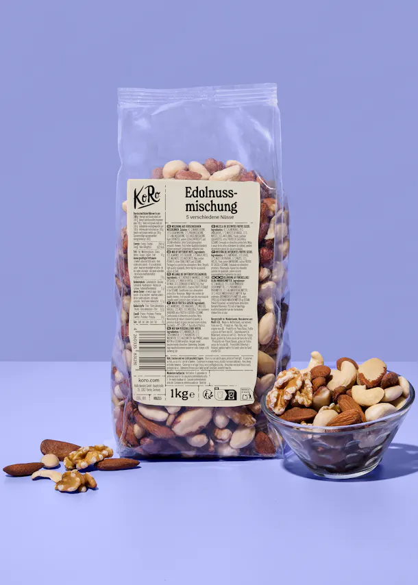 eine klare plastiktüte mit einer 1kg ko ro edelnuss-mischung aus 5 verschiedenen nüssen steht auf einem lila hintergrund, daneben eine glasschale mit nüssen und einige verstreute nüsse.