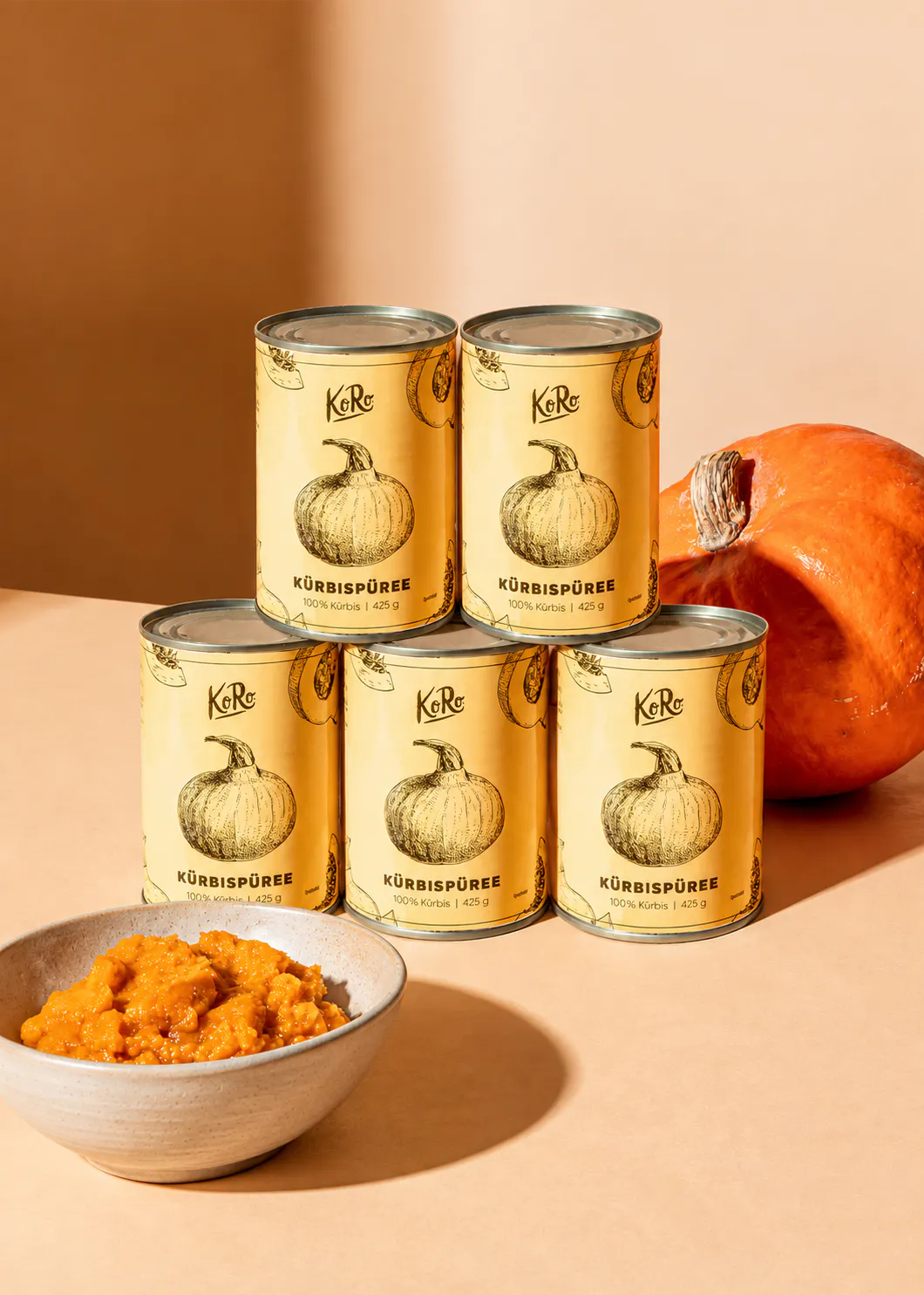 cinque barattoli di purea di zucca koro disposti a piramide accanto a una zucca intera e una ciotola di purea fresca.