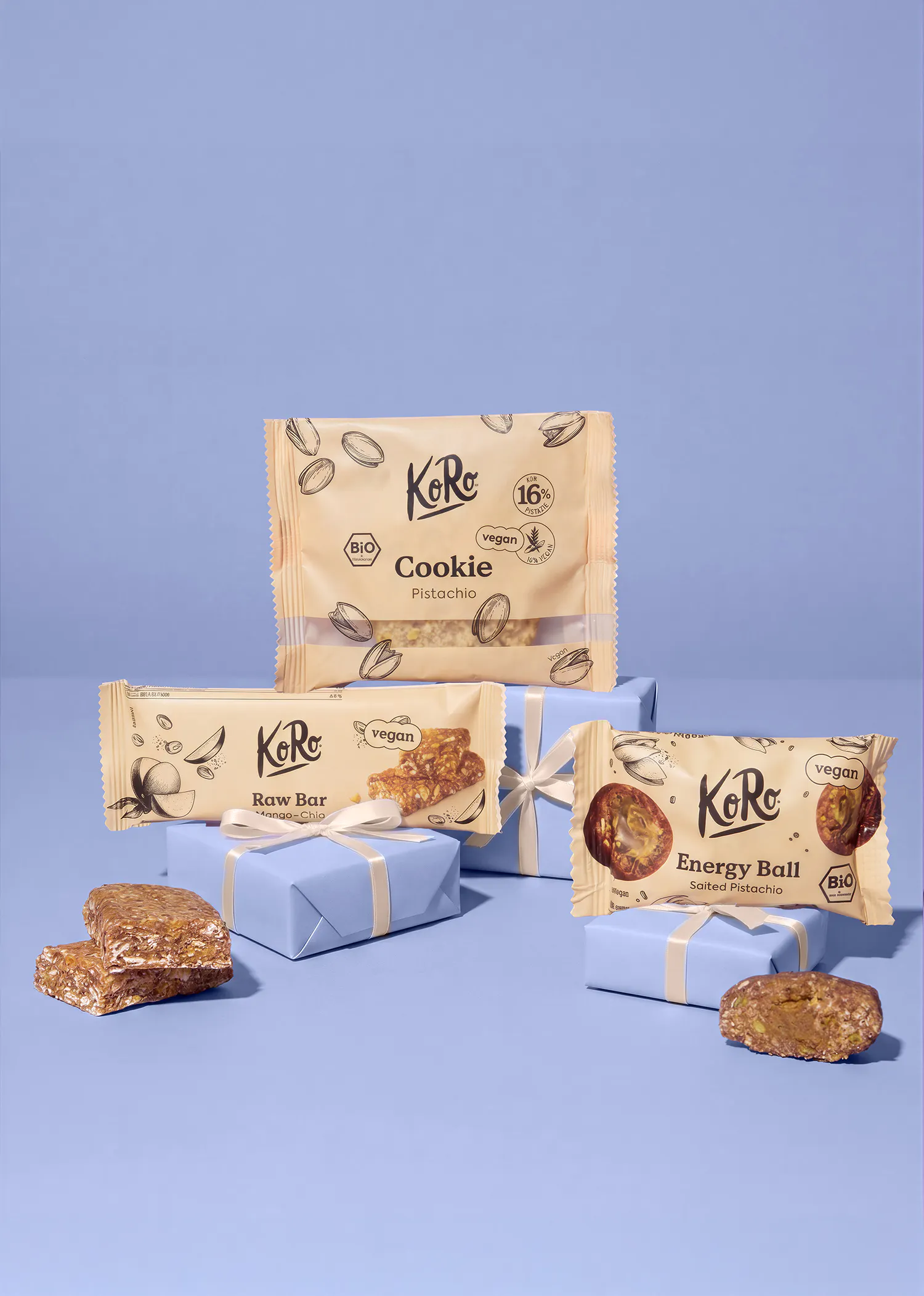trois snacks végétaliens koro (un cookie à la pistache, une barre crue mangue-chia et une boule d'énergie pistache salée) sont présentés sur des boîtes cadeaux bleu clair, certains ouverts pour montrer l'intérieur, sur un fond bleu.