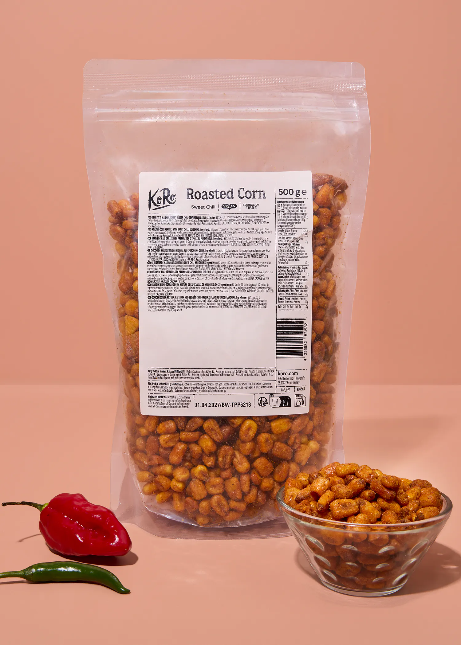 sachet de maïs grillé saveur piment doux de la marque koro posé sur une surface rose à côté d'un bol rempli de maïs et de deux piments frais.
