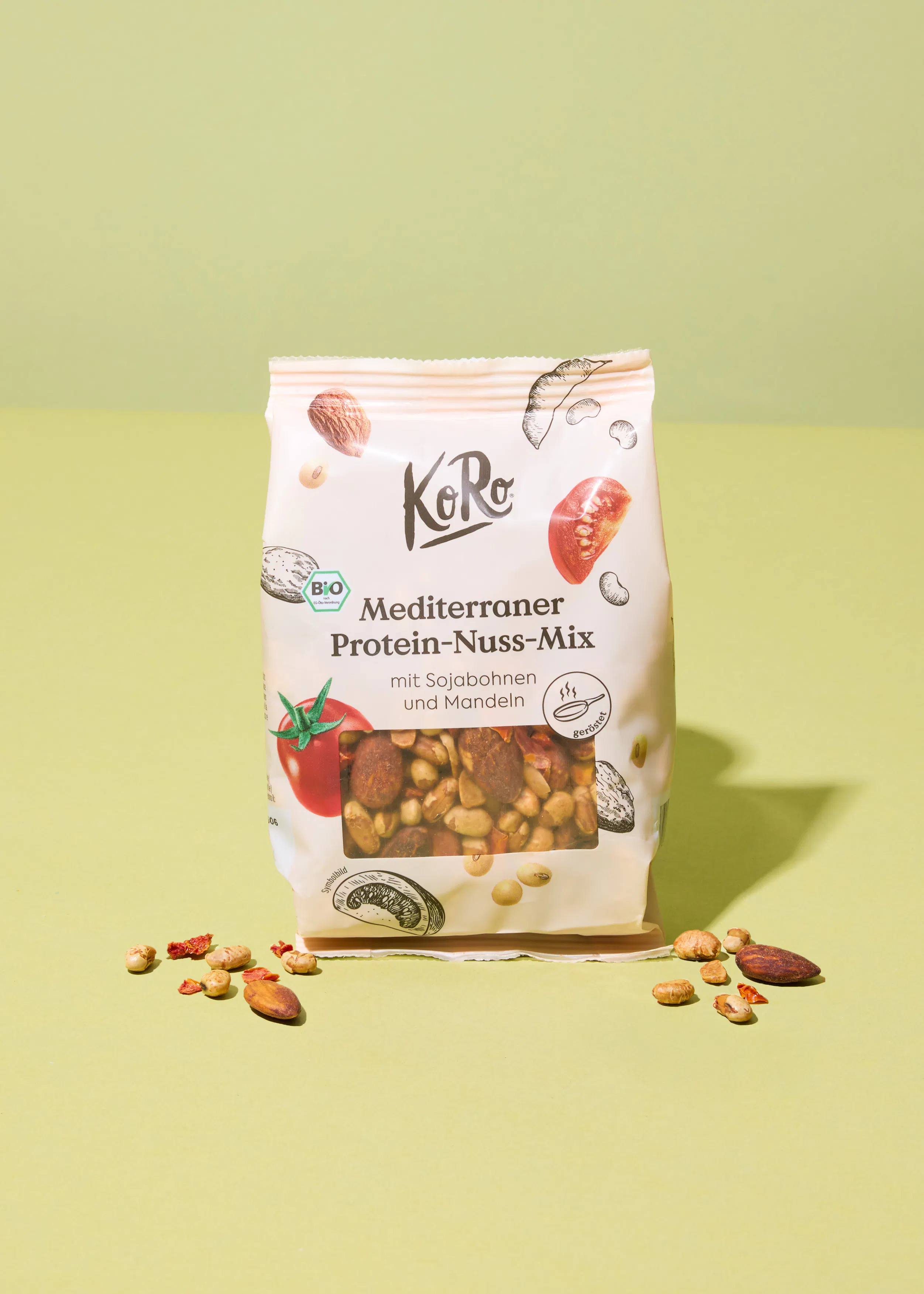 una bolsa blanca de KoRo mediterraner protein-nuss-mix con soja y almendras, con algunas nueces y tomates secos esparcidos sobre un fondo verde claro.