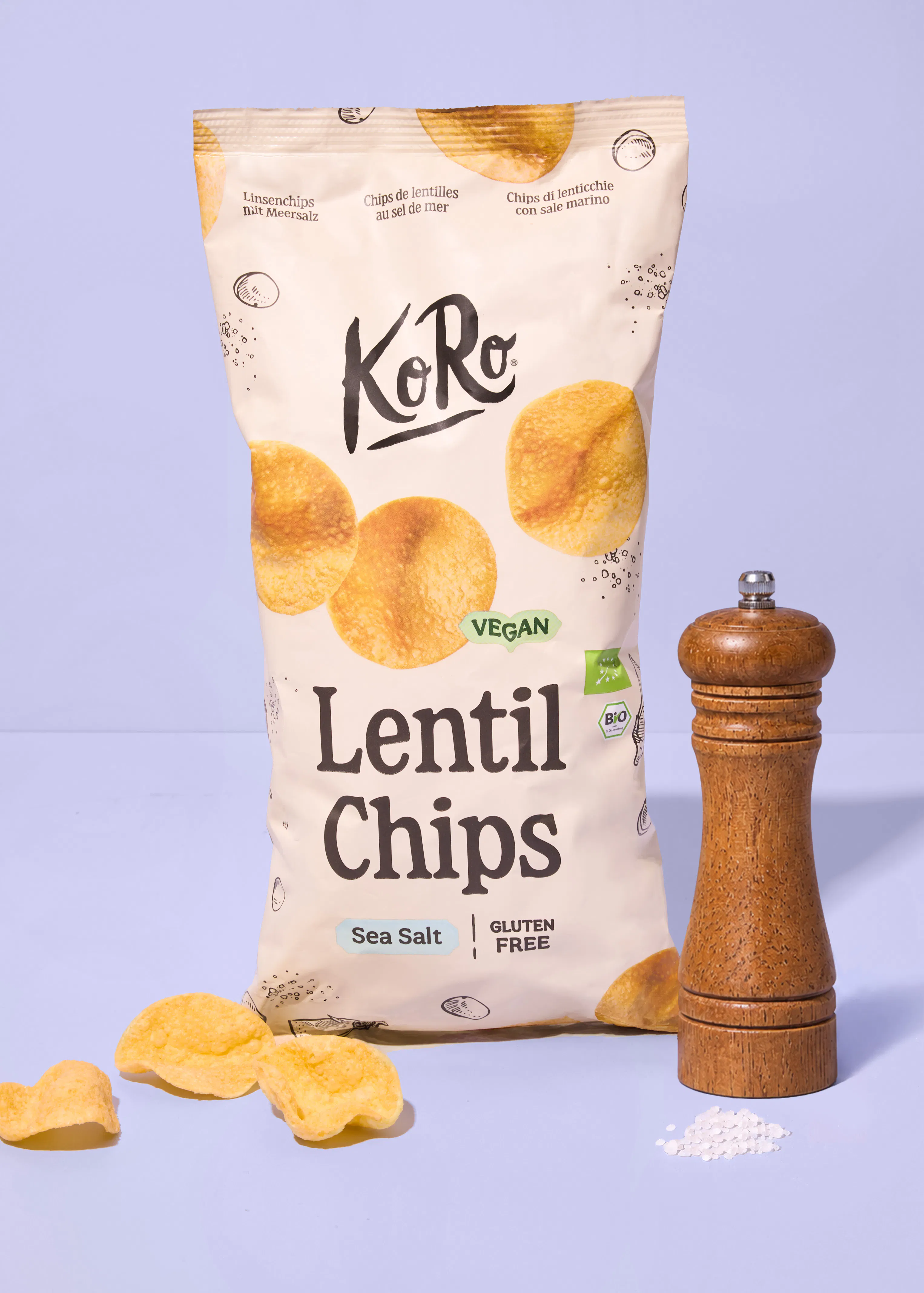 a bag of koro brand sea salt lentil chips stands next to a wooden pepper mill with a few loose chips and salt grains on a light purple surface.