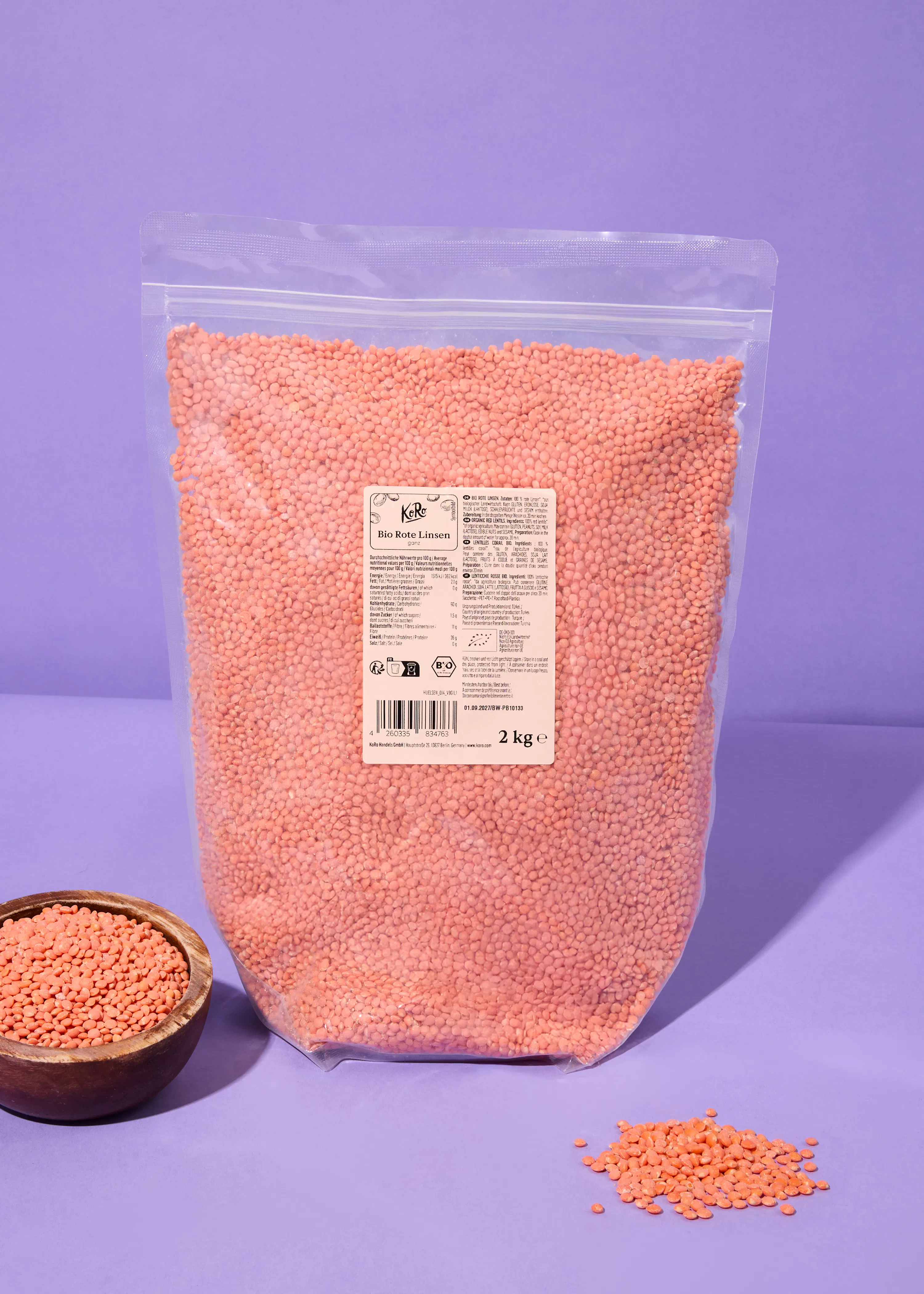 eine große durchsichtige tüte mit 2 kg kero bio roten linsen steht auf einem lila untergrund, daneben eine holzschale mit linsen und ein kleiner haufen linsen.
