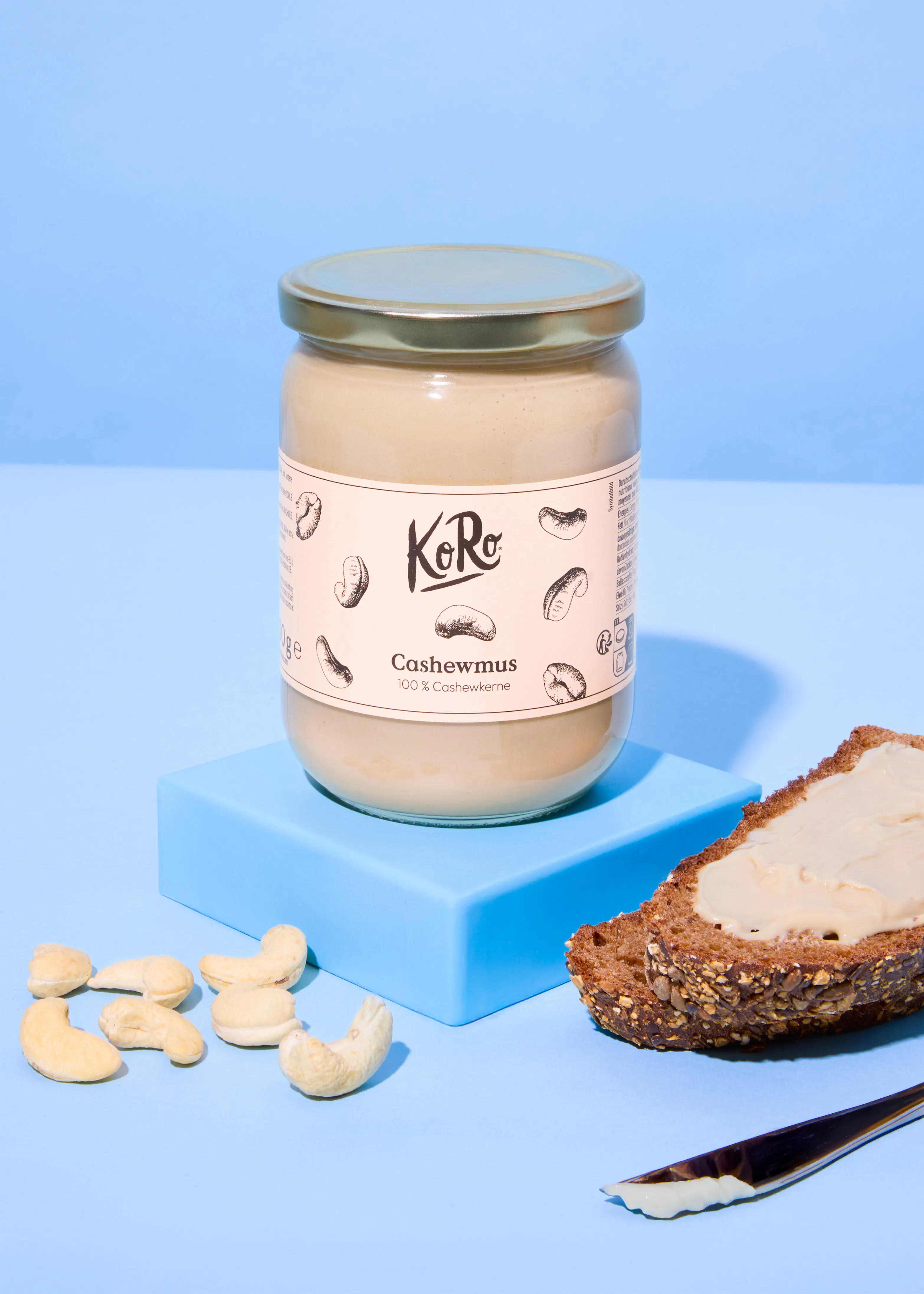 un pot de beurre de cajou koro est posé sur un bloc bleu, avec des noix de cajou éparpillées, une tranche de pain tartinée de beurre de cajou et un couteau sur une surface bleue.