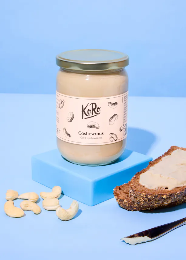 a jar of koro cashew butter sits on a blue block next to loose cashews and a slice of bread with cashew butter spread on it, with a knife.