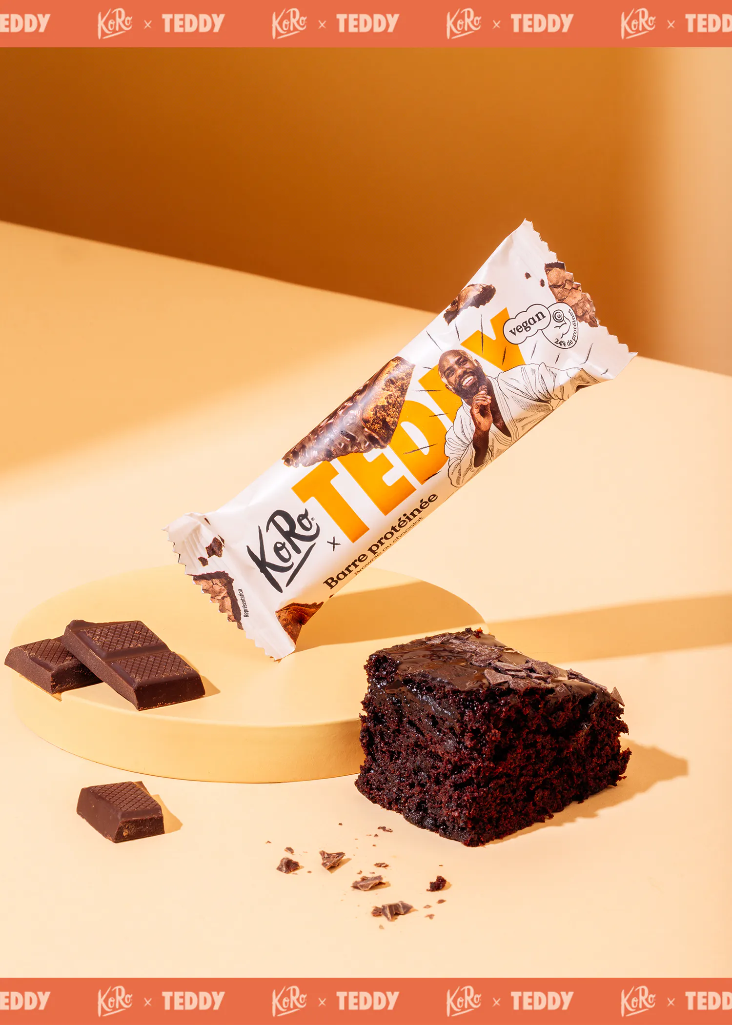 une barre protéinée koro x teddy végane saveur brownie au chocolat, un morceau de brownie et des carrés de chocolat sont présentés sur un fond orange avec une plateforme jaune.