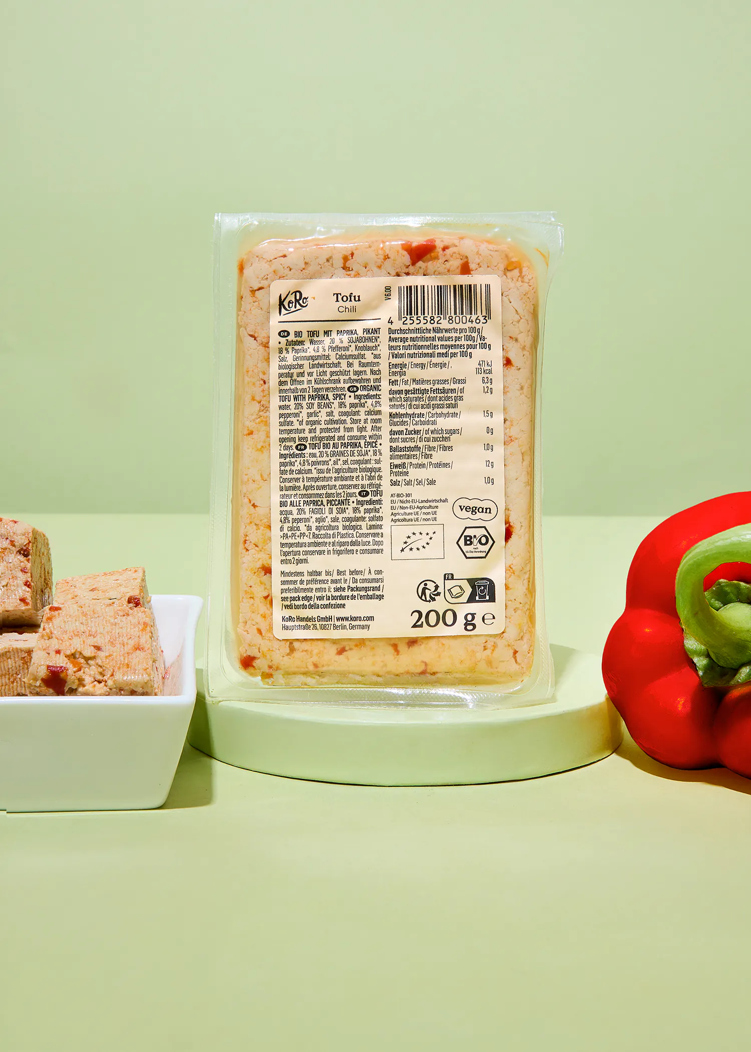 a partially unwrapped package of koro tofu chili sits on a light green surface next to a red bell pepper and a white dish with tofu cubes.