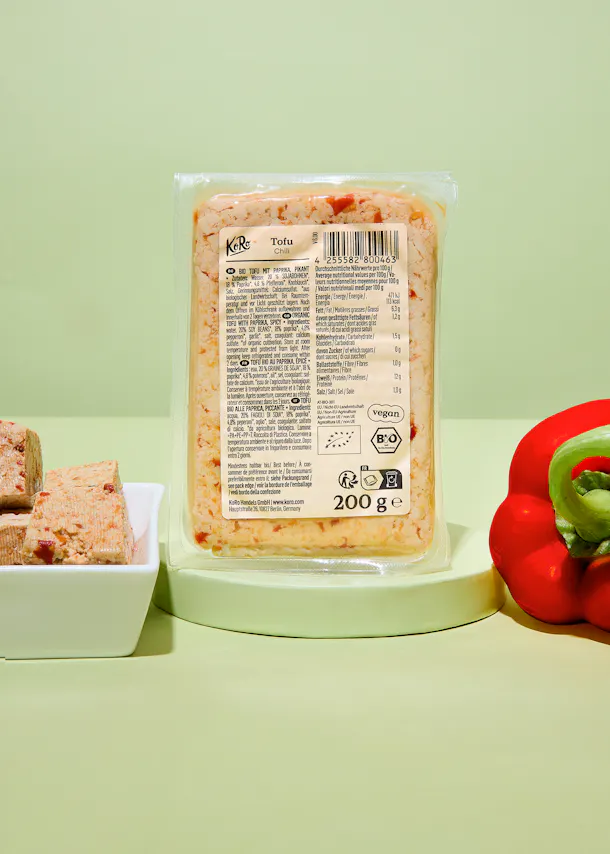 a partially unwrapped package of koro tofu chili sits on a light green surface next to a red bell pepper and a white dish with tofu cubes.
