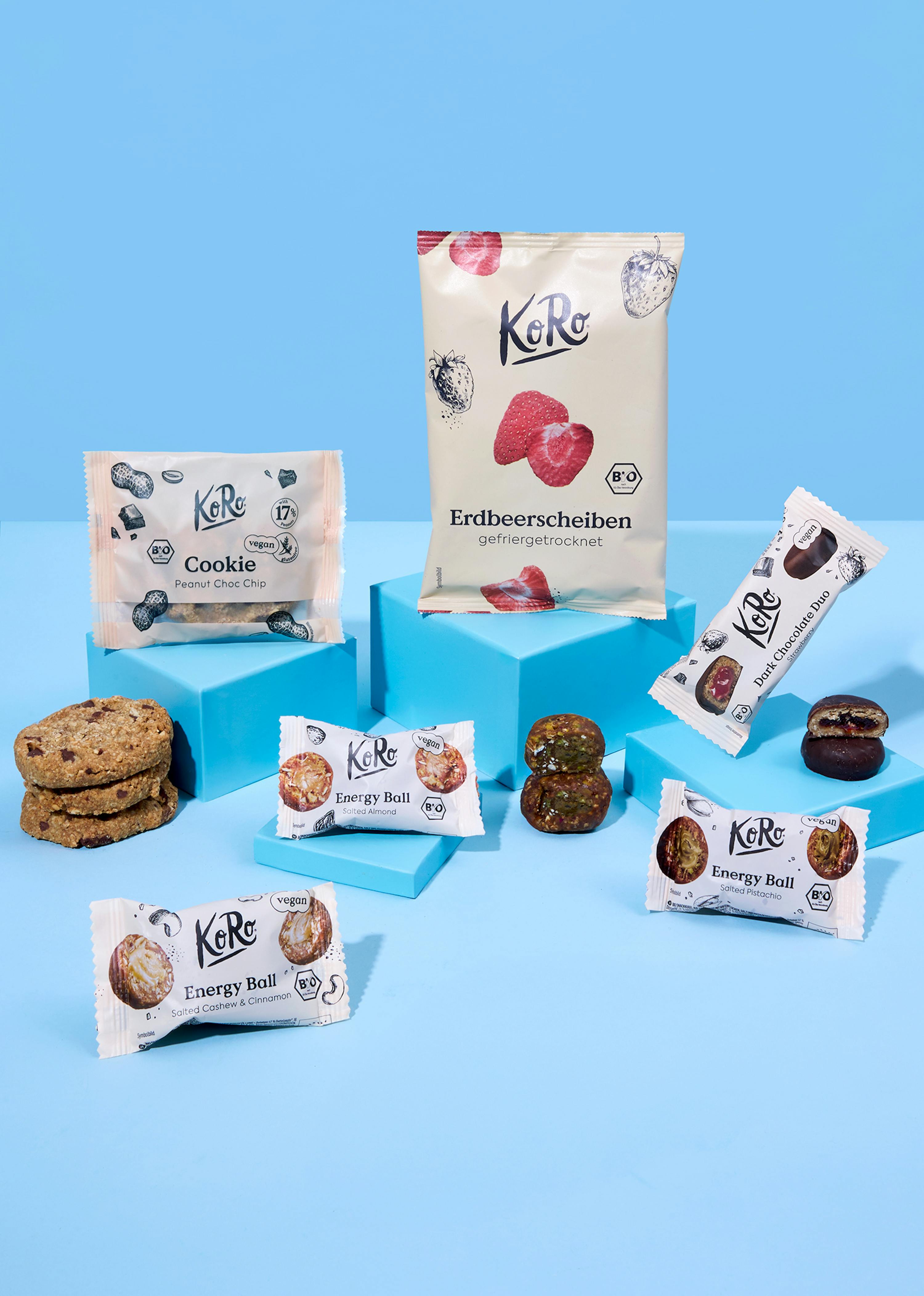 una varietà di snack ko ro, inclusi biscotti, palline energetiche, fette di fragola liofilizzate e barrette di cioccolato, disposti su blocchi blu su uno sfondo azzurro.