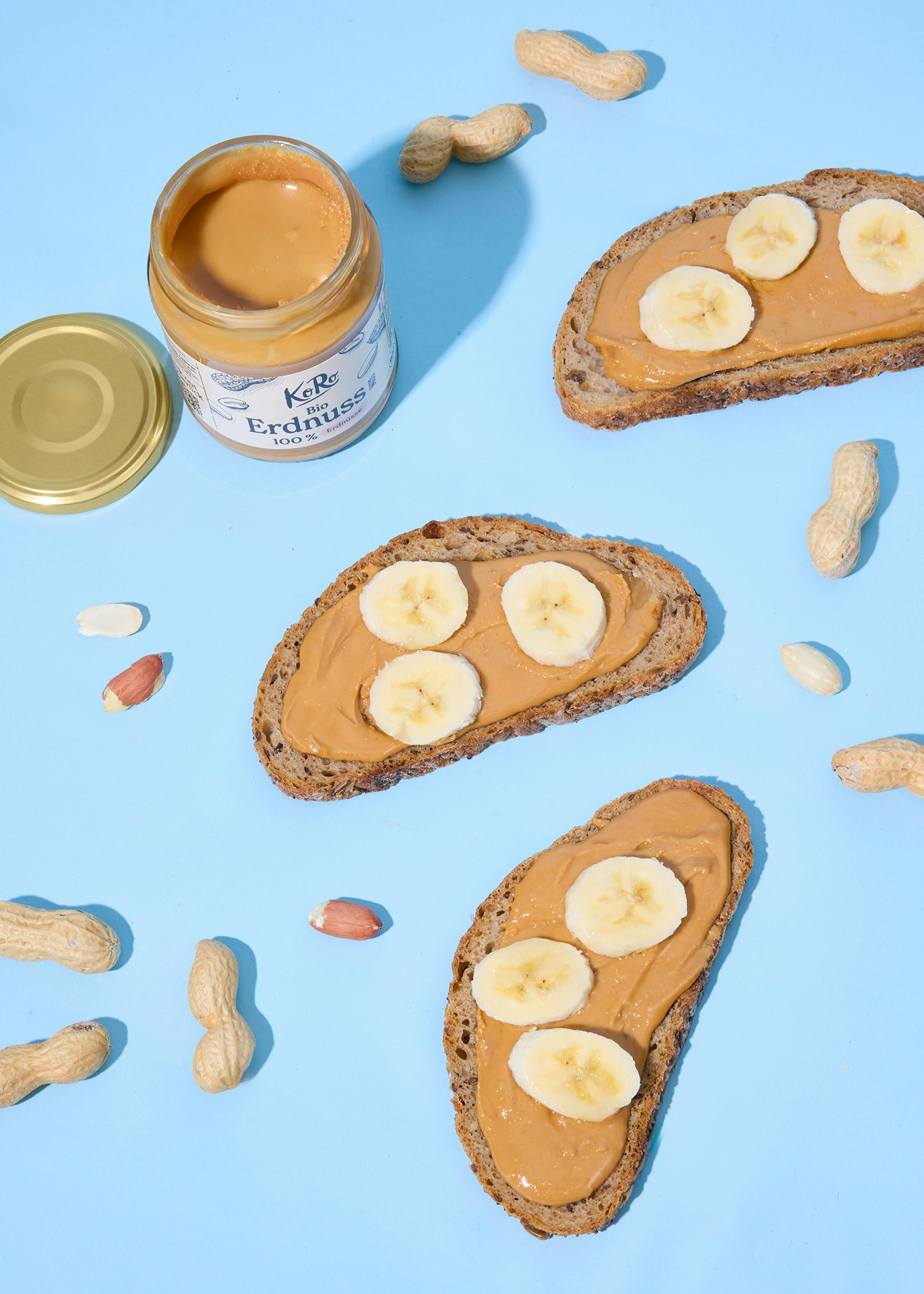 a jar of peanut butter, three slices of peanut butter toast with banana, and scattered peanuts on a blue background.