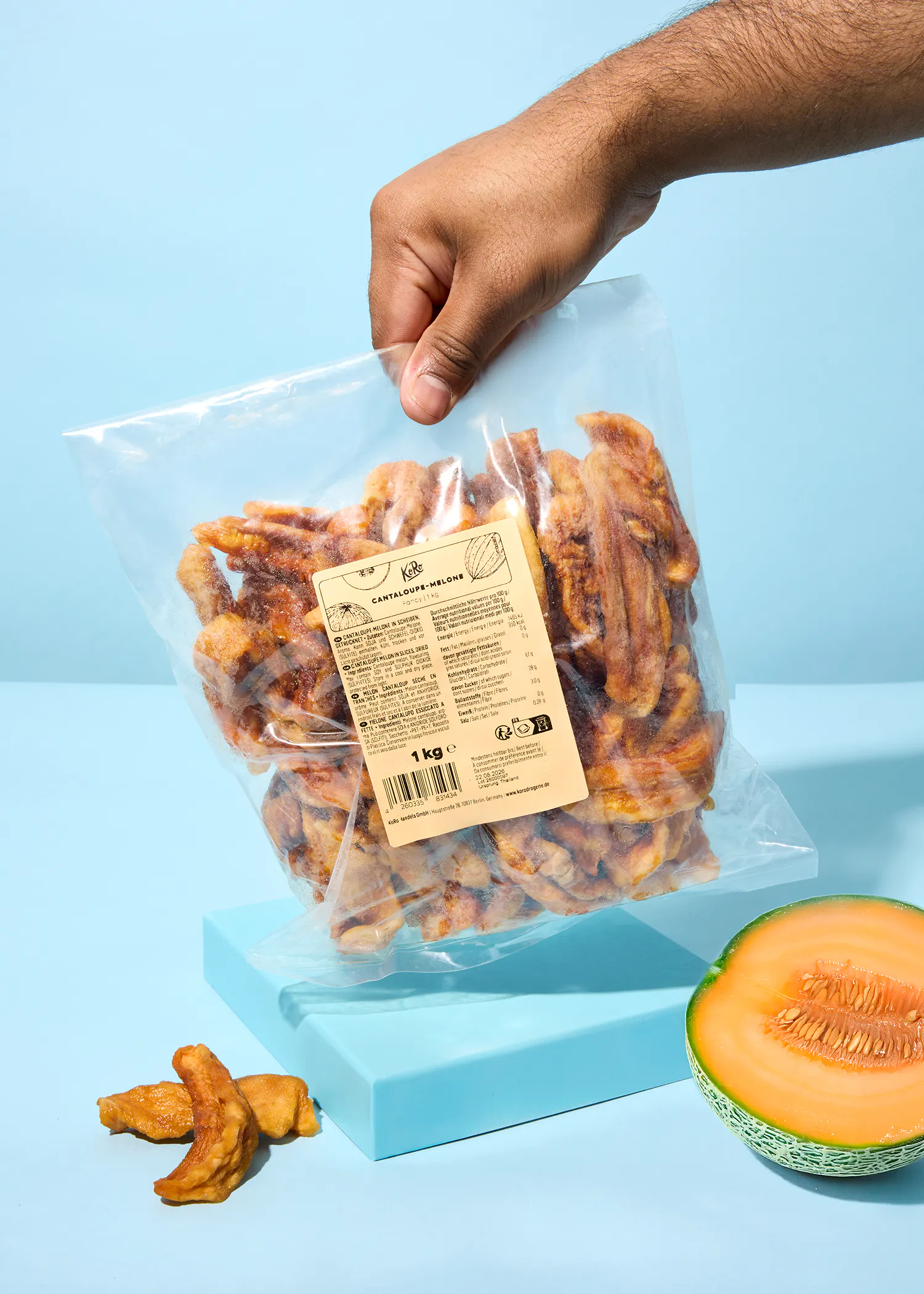 a hand holds a clear bag of dried cantaloupe slices above a blue surface, next to a fresh cantaloupe half and a few loose dried slices.