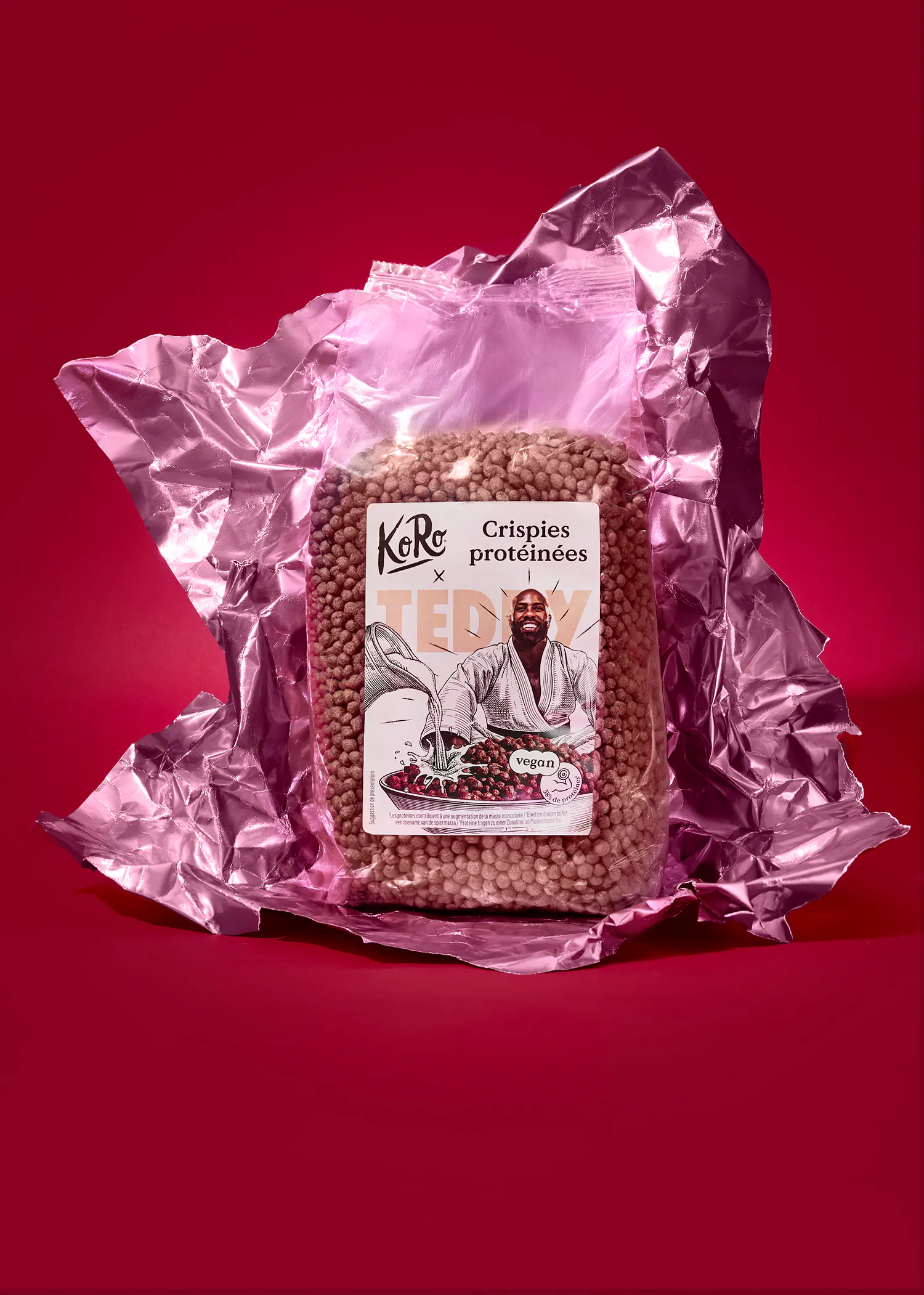 un sac transparent de crispies protéinées koro x ted by, avec une étiquette montrant un homme souriant en robe versant des baies et le logo vegan, posé sur un fond rose métallique froissé et rouge uni.