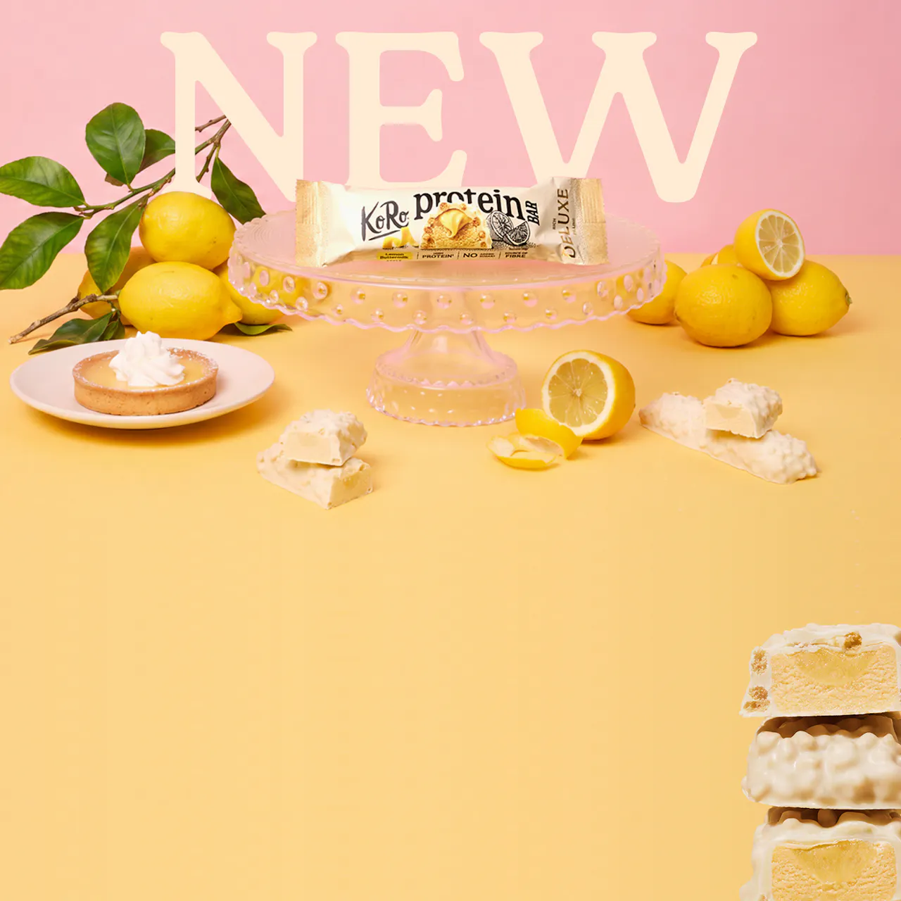 ein koro proteinriegel in der geschmacksrichtung zitrone-buttermilch wird auf einer gläsernen etagere präsentiert, umgeben von frischen zitronen und einem kleinen zitronentörtchen vor einem pastellfarbenen hintergrund mit dem schriftzug new.