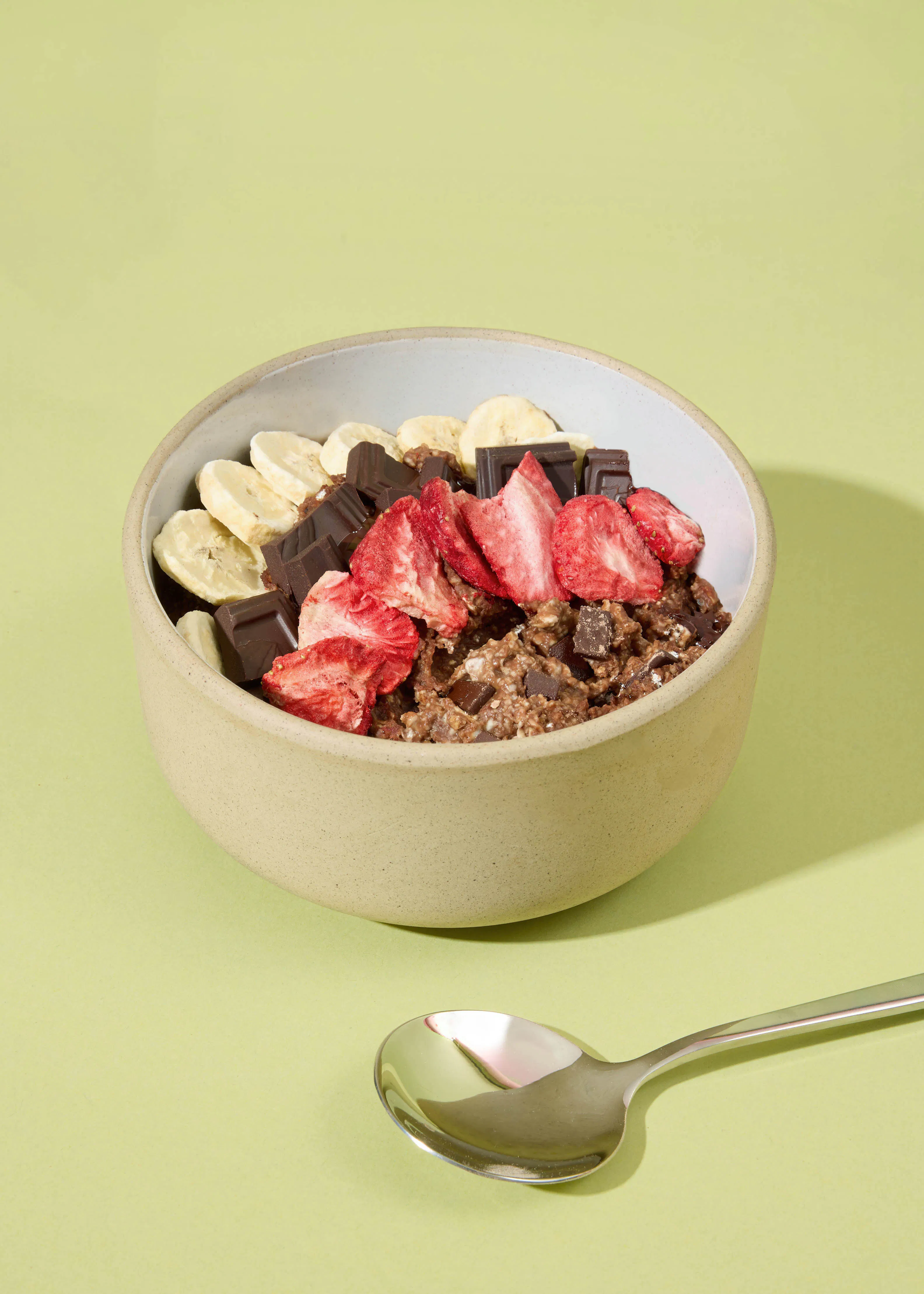 a bowl of cereal with bananas chocolate and strawberries next to a spoon