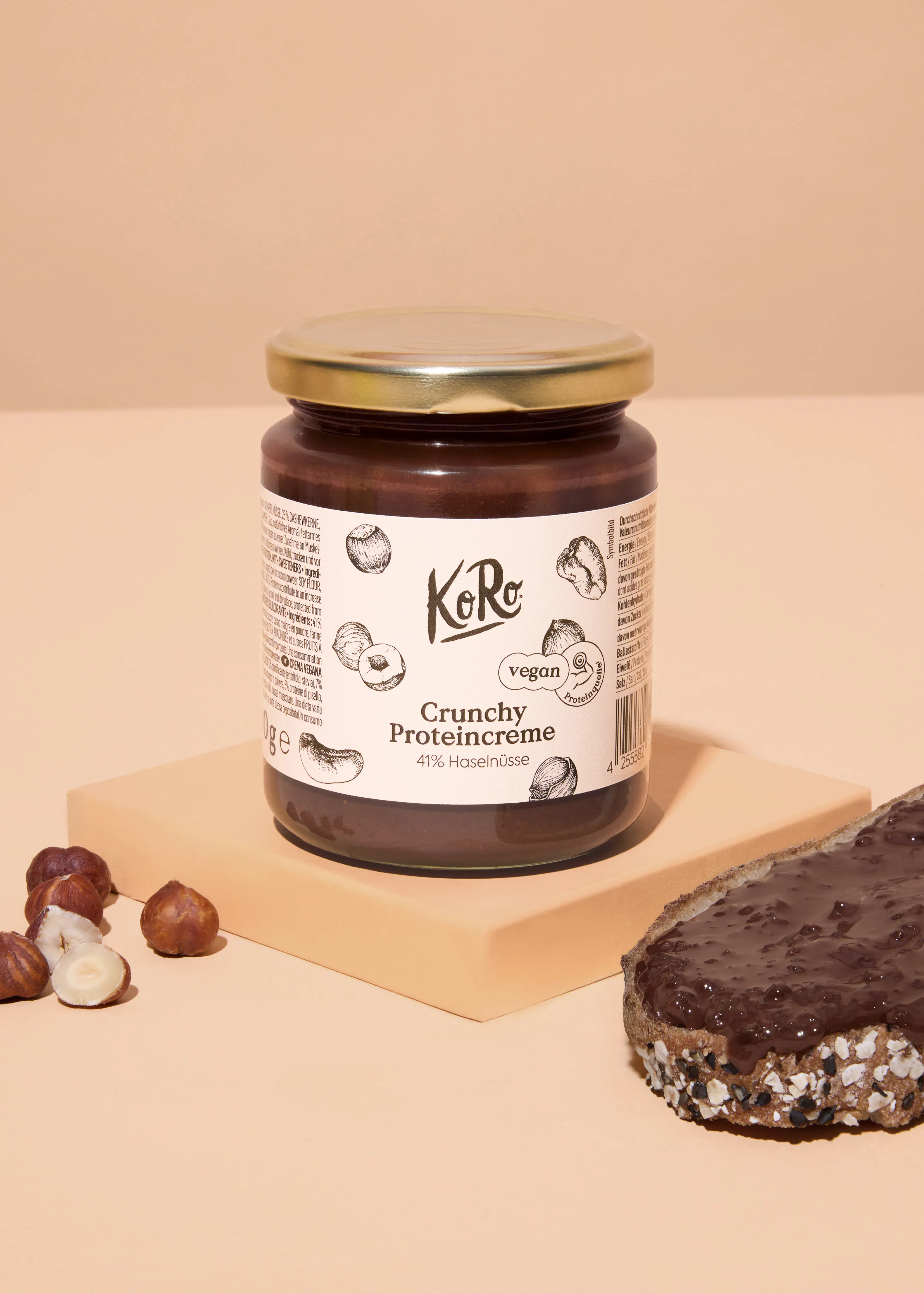 un pot de crème protéinée croquante aux noisettes koro, des noisettes et une tranche de pain tartinée de cette crème sont présentés sur un fond beige.