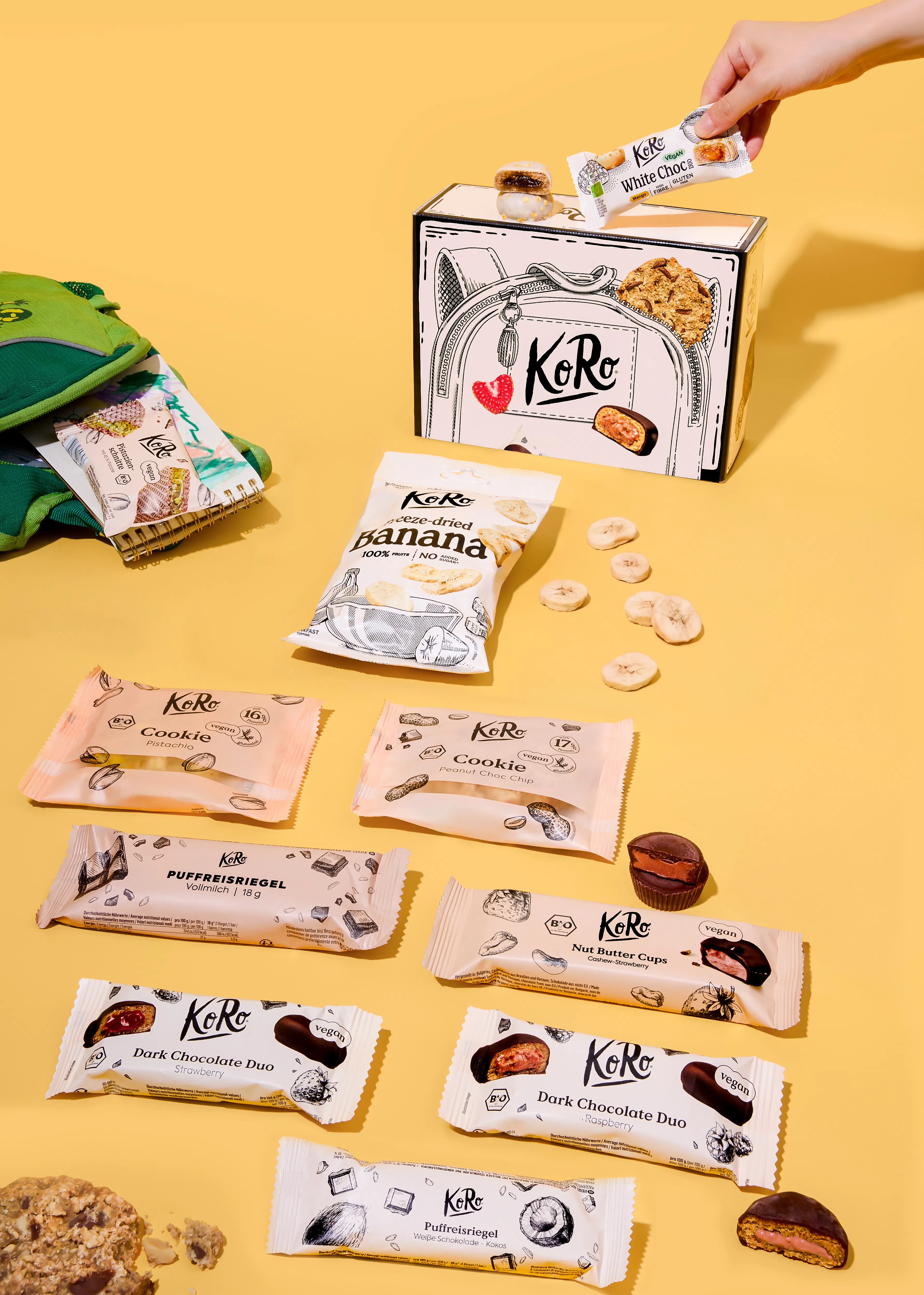 eine anordnung verschiedener koro-snacks wie riegel, kekse und gefriergetrocknete bananenscheiben auf einem gelben hintergrund.