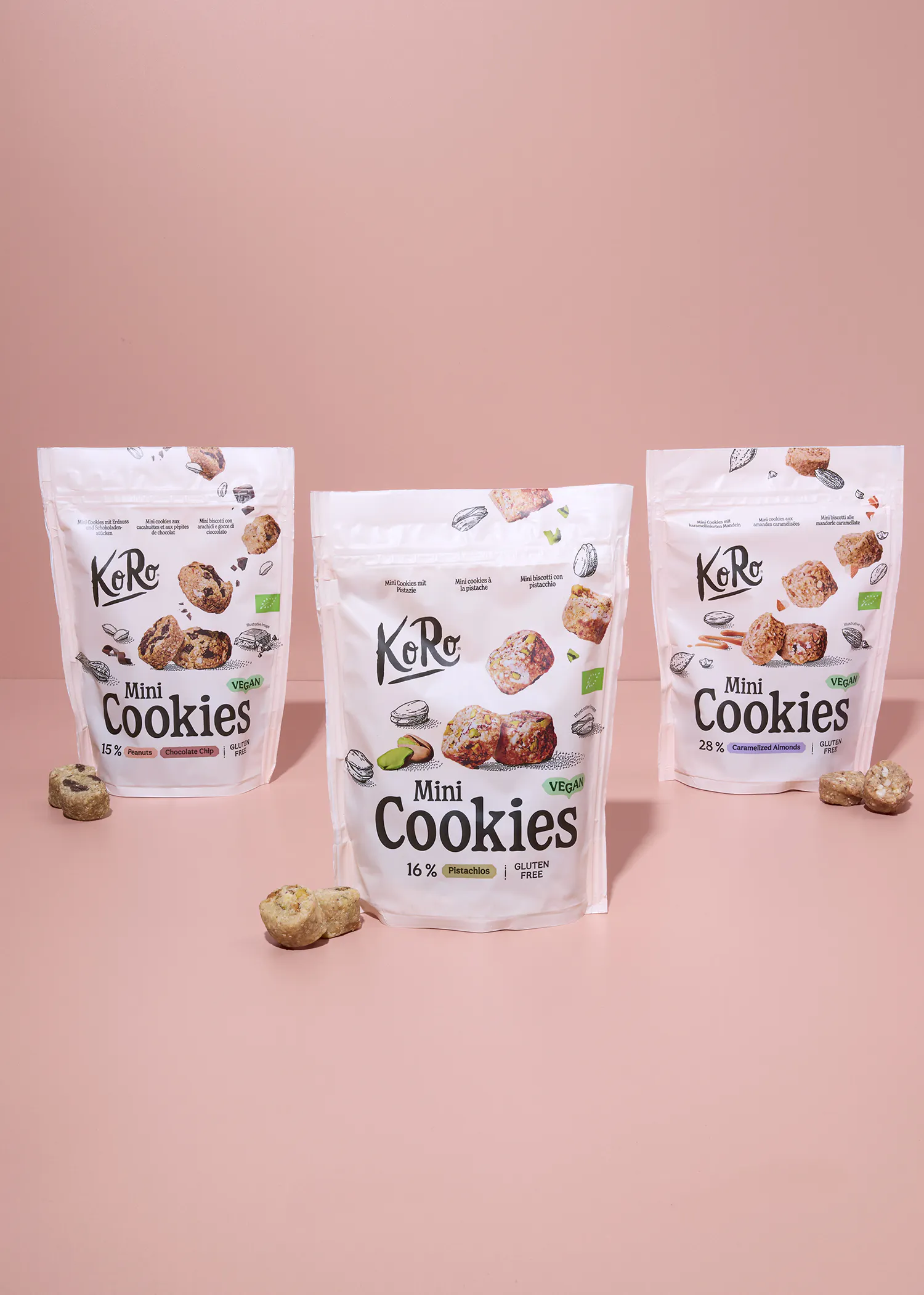 drei weiße beutel mit koro mini cookies in den sorten erdnuss-schokoladenstückchen, pistazie und karamellisierte mandeln stehen auf einer rosa oberfläche, umgeben von einigen verstreuten keksen.