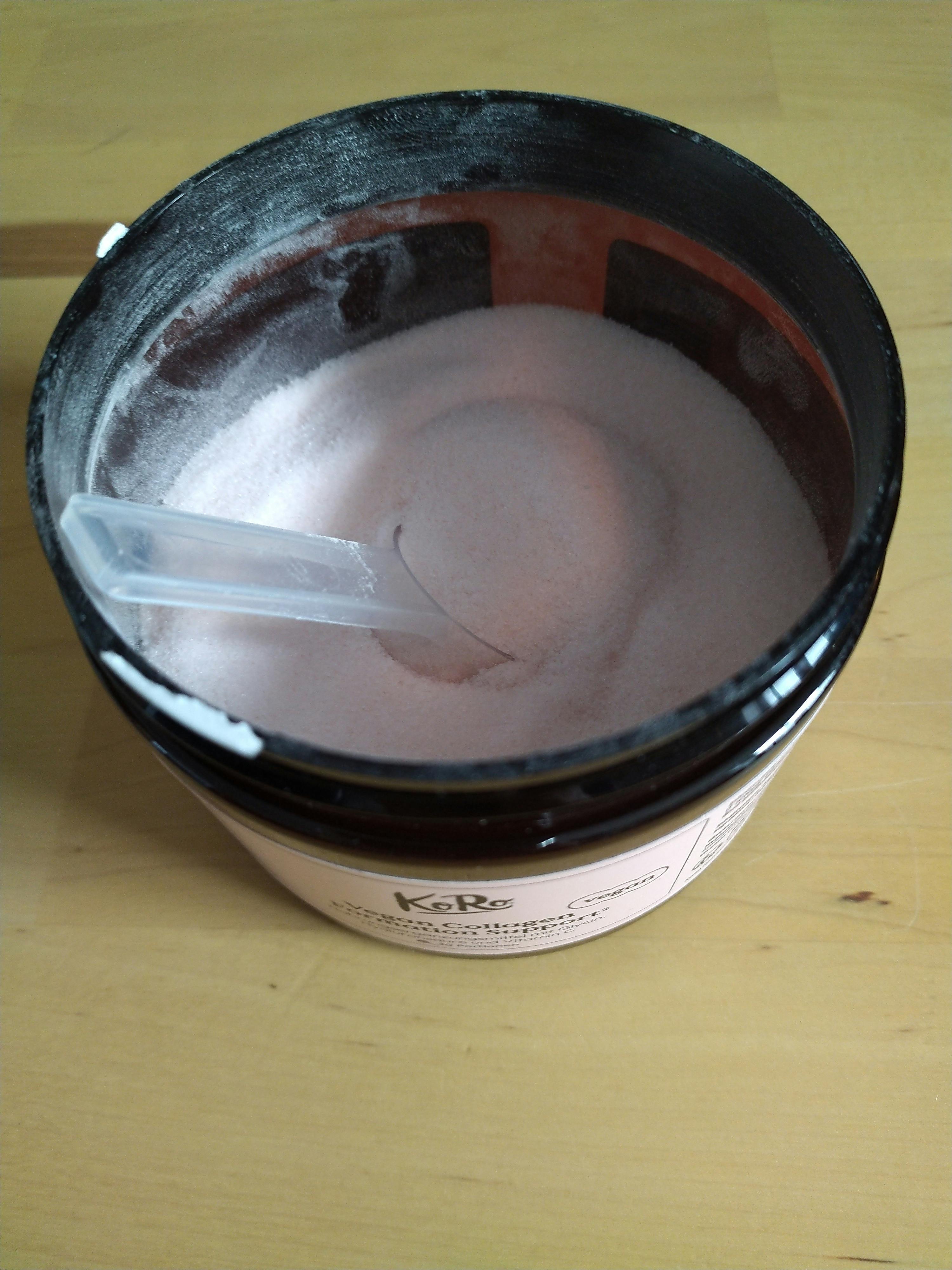 an open black koro jar filled with light pink vegan collagen support powder and a clear scoop, sitting on a wooden surface.