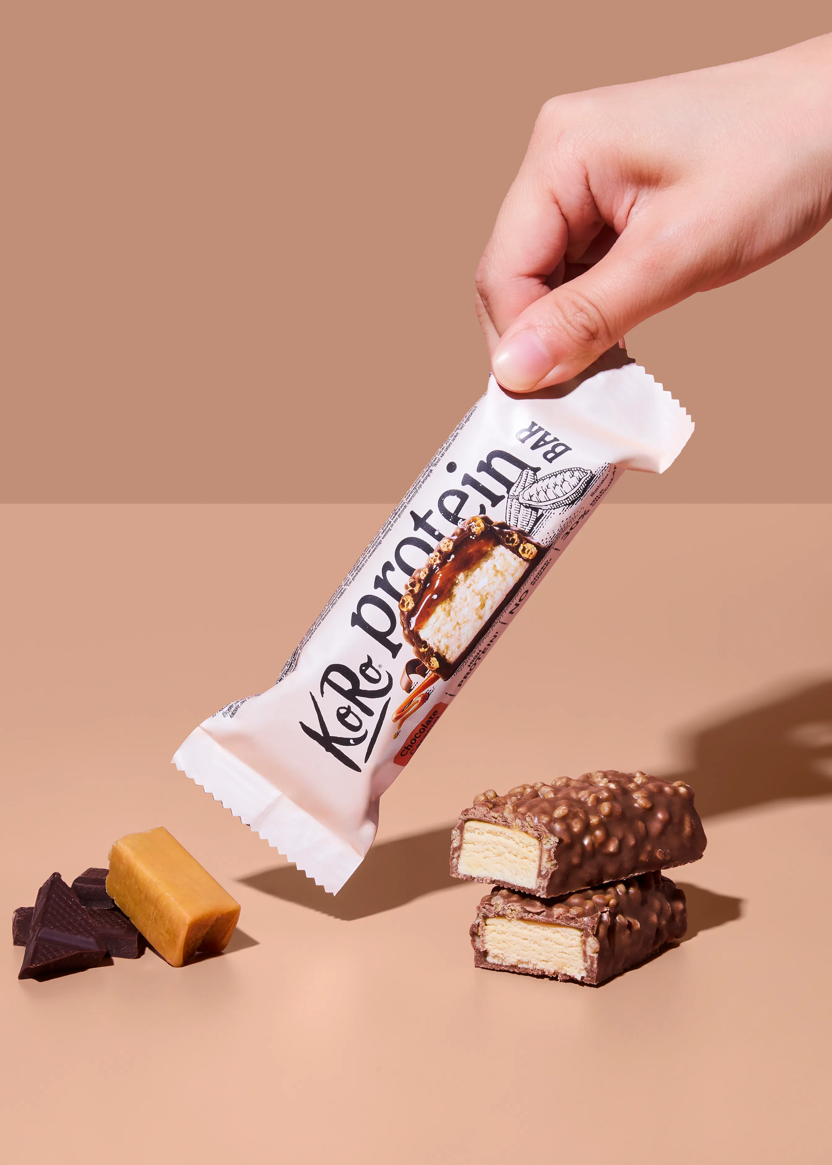 eine hand hält eine koro proteinriegelverpackung, daneben liegen zwei halbierte schokoriegel mit weißer füllung, karamell und dunkle schokoladenstücke.