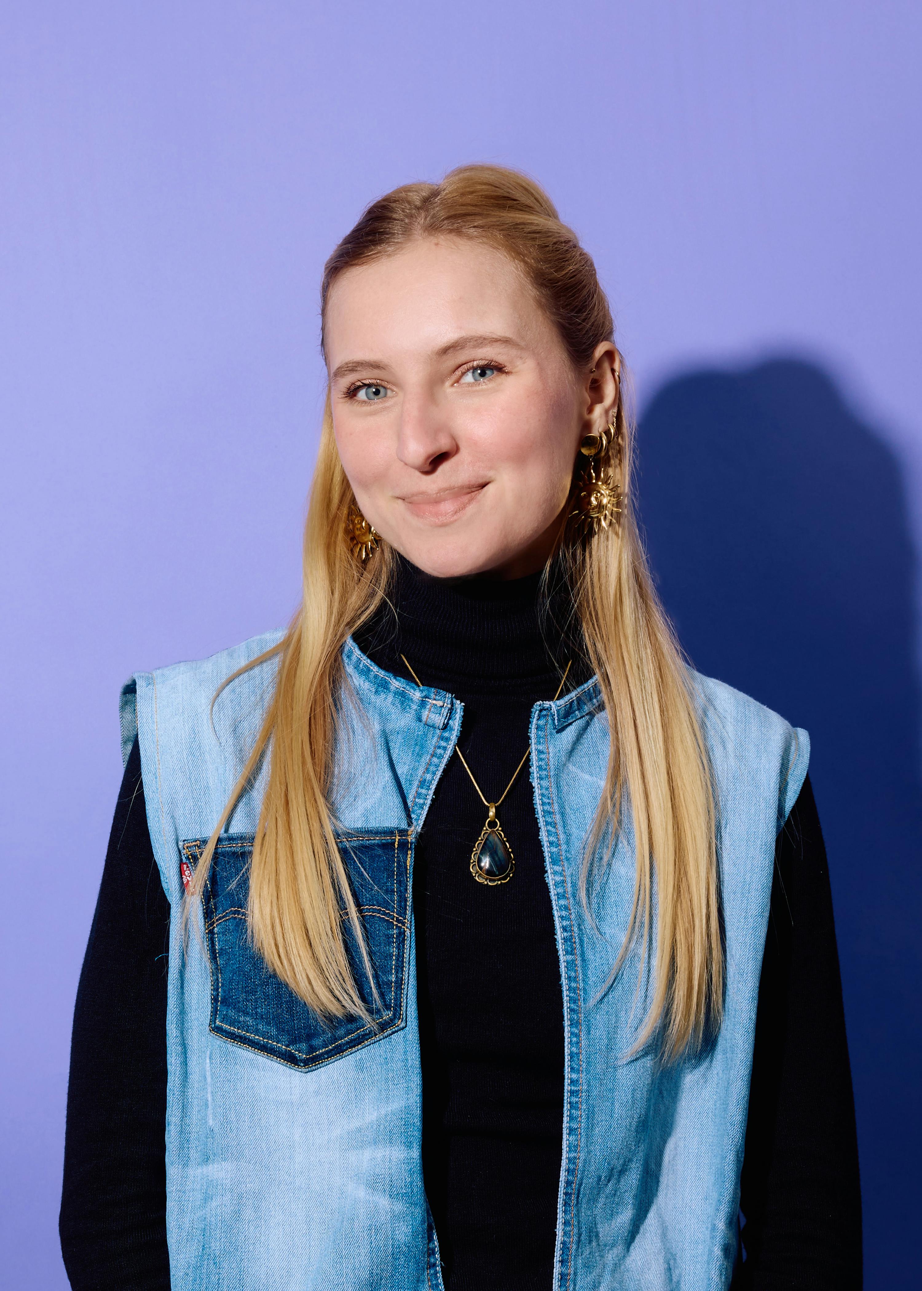 eine junge frau mit langen blonden haaren, blauen augen und einem leichten lächeln trägt einen schwarzen rollkragenpullover, eine jeansweste, goldene ohrringe und eine goldene halskette mit einem dunklen anhänger vor einem lila hintergrund.